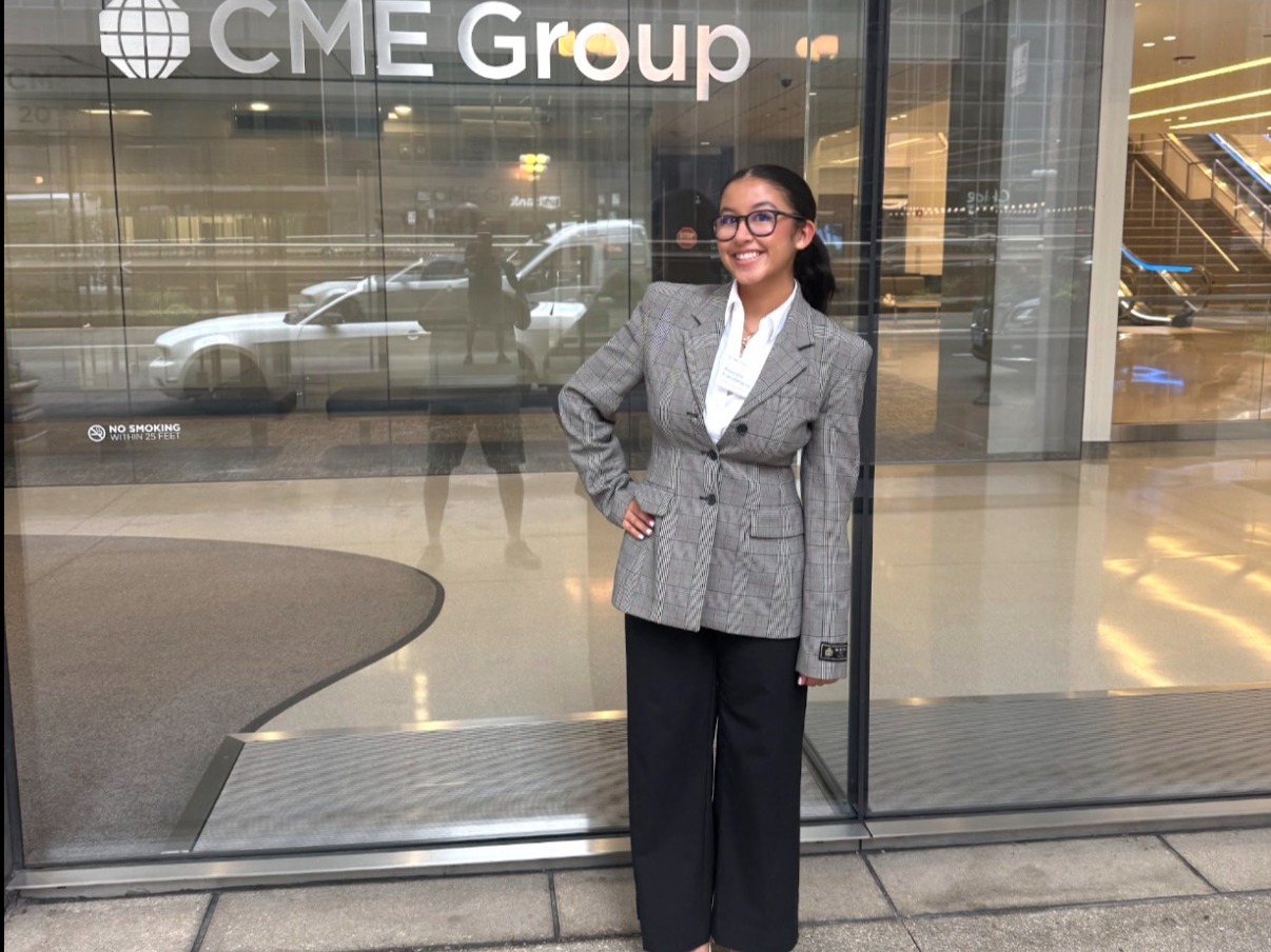 A smiling young woman in glasses, a gray plaid blazer, white shirt, black wide-leg trousers, and white heels poses with a hand on her hip outside the glass entrance of a building with a CME Group sign.