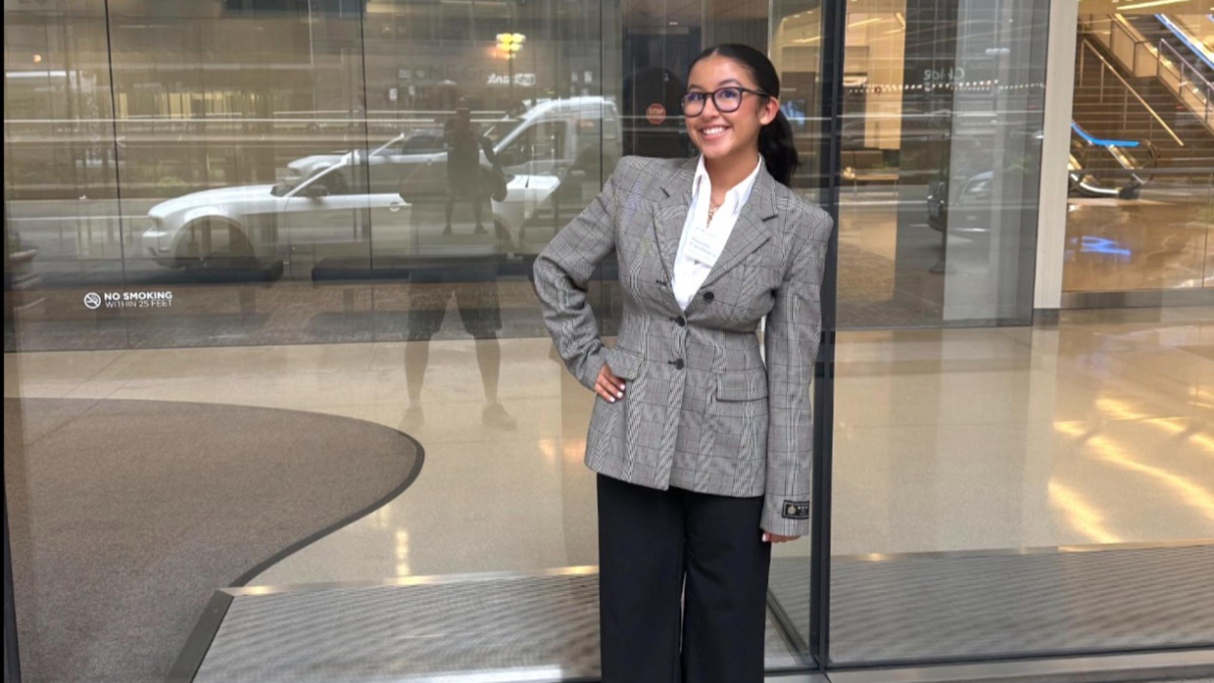 A smiling young woman in glasses, a gray plaid blazer, white shirt, black wide-leg trousers, and white heels poses with a hand on her hip outside the glass entrance of a building with a CME Group sign.