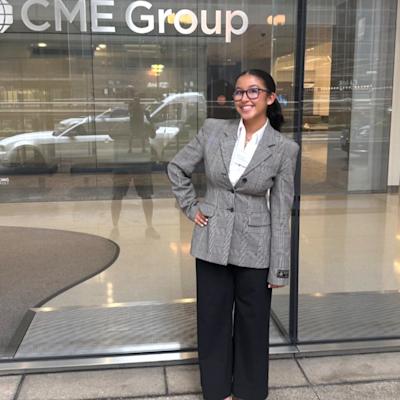 A smiling young woman in glasses, a gray plaid blazer, white shirt, black wide-leg trousers, and white heels poses with a hand on her hip outside the glass entrance of a building with a CME Group sign.