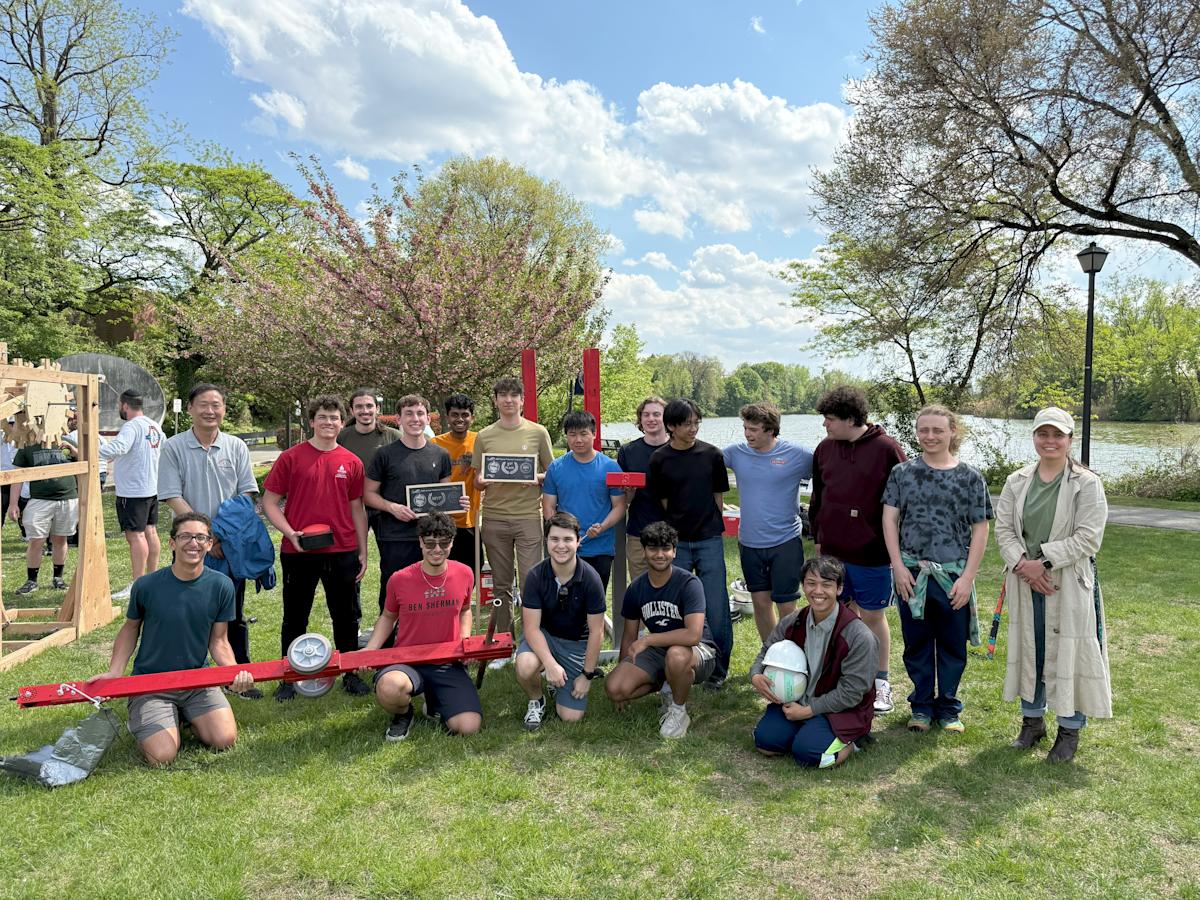 The team of students sit and stand in two rows smiling at the camera, behind the arm of their catapult.