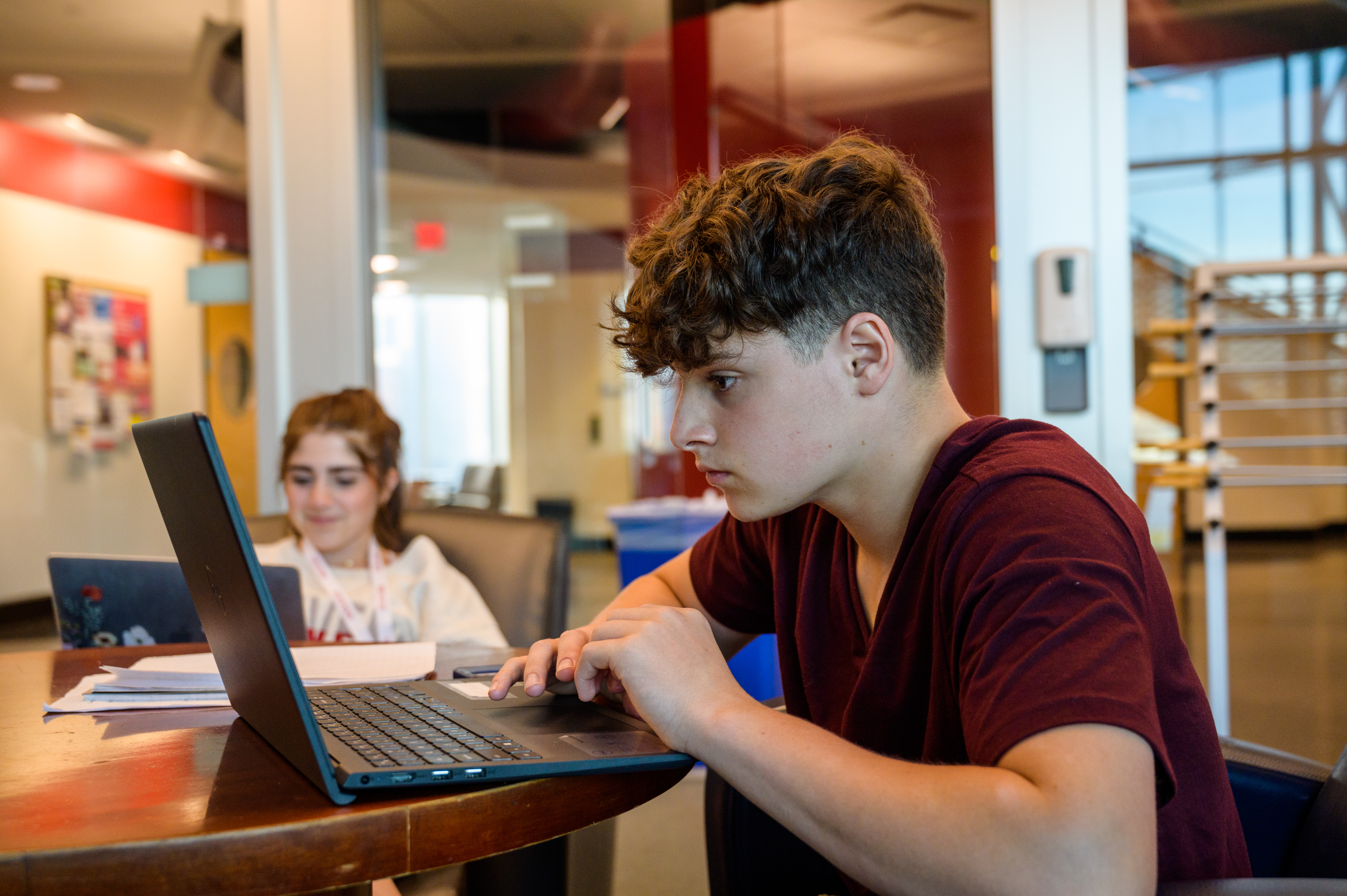 Pre-College student looks intently at his laptop while anopther student smiles in the background.