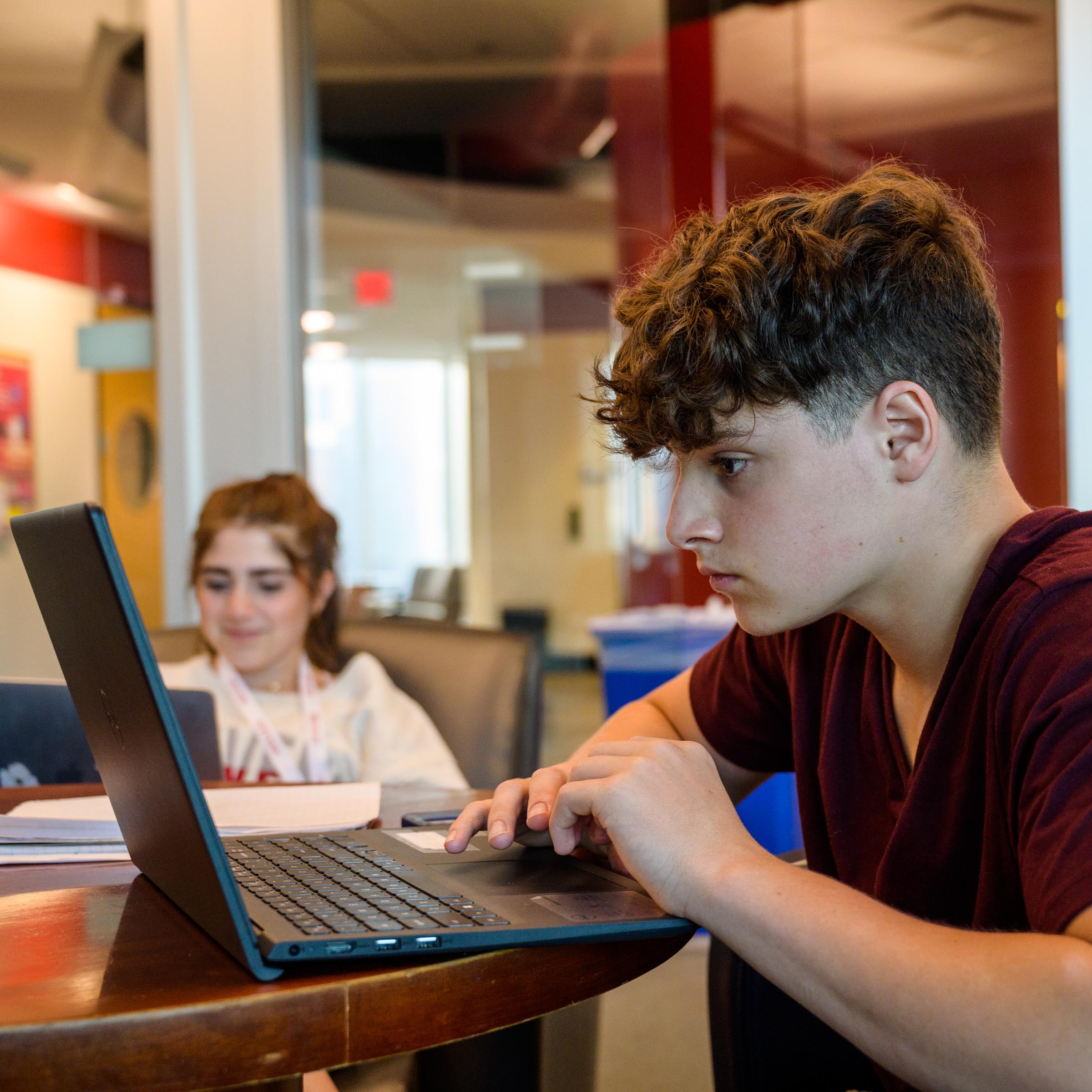 Pre-College student looks intently at his laptop while anopther student smiles in the background.