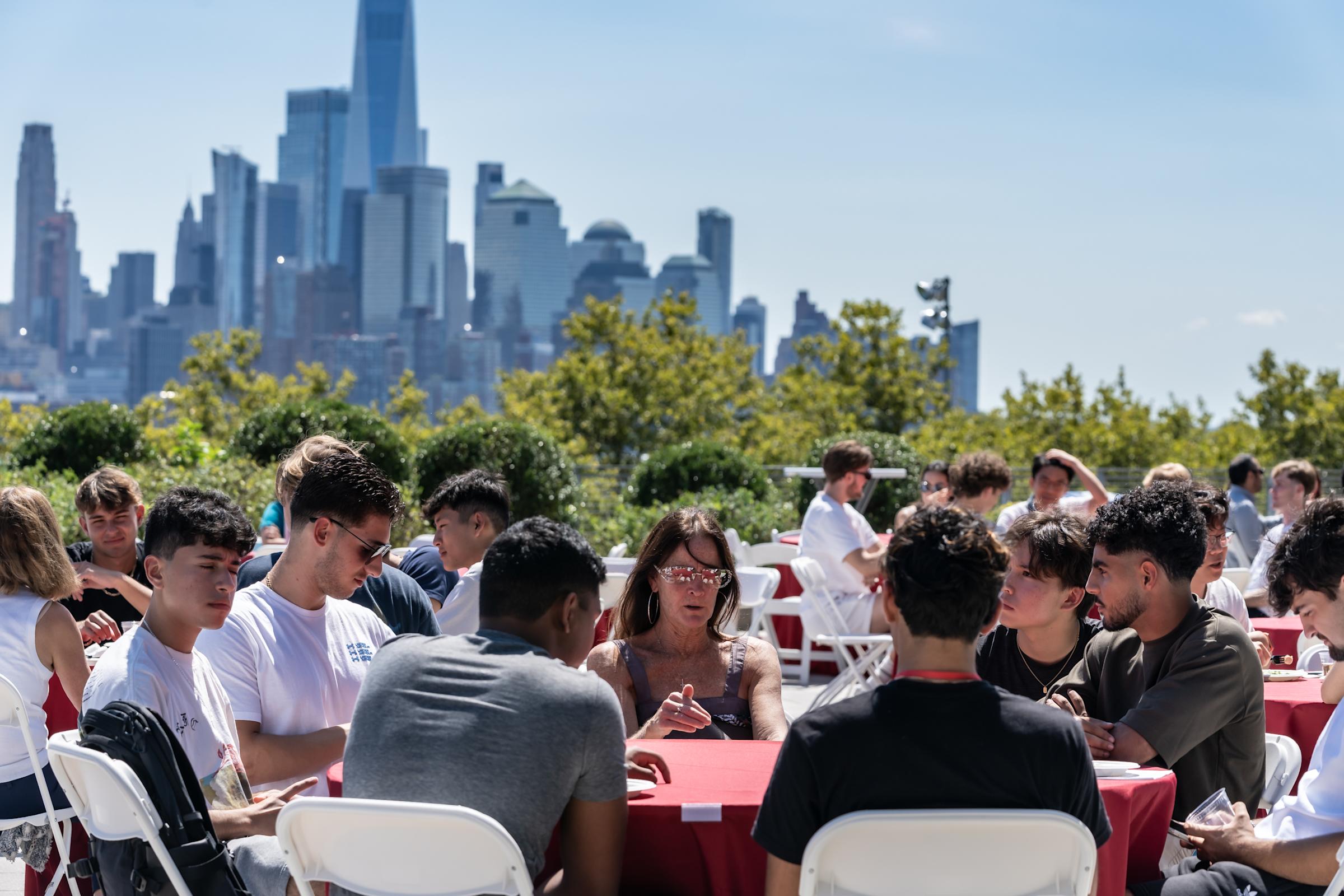 Students gathered around red tables for outdoor dining event on a terrace, with Manhattan skyline visible in background on clear day.