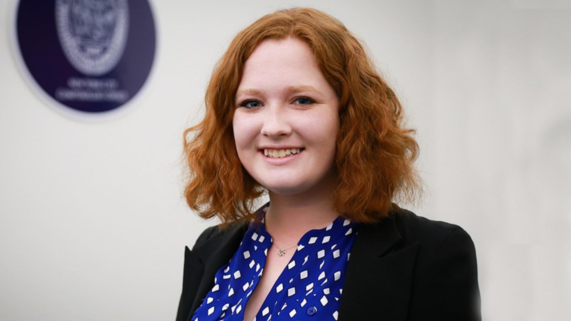 Victoria Piskarev in a blue dress and black blazer before the official seal of New York City government.