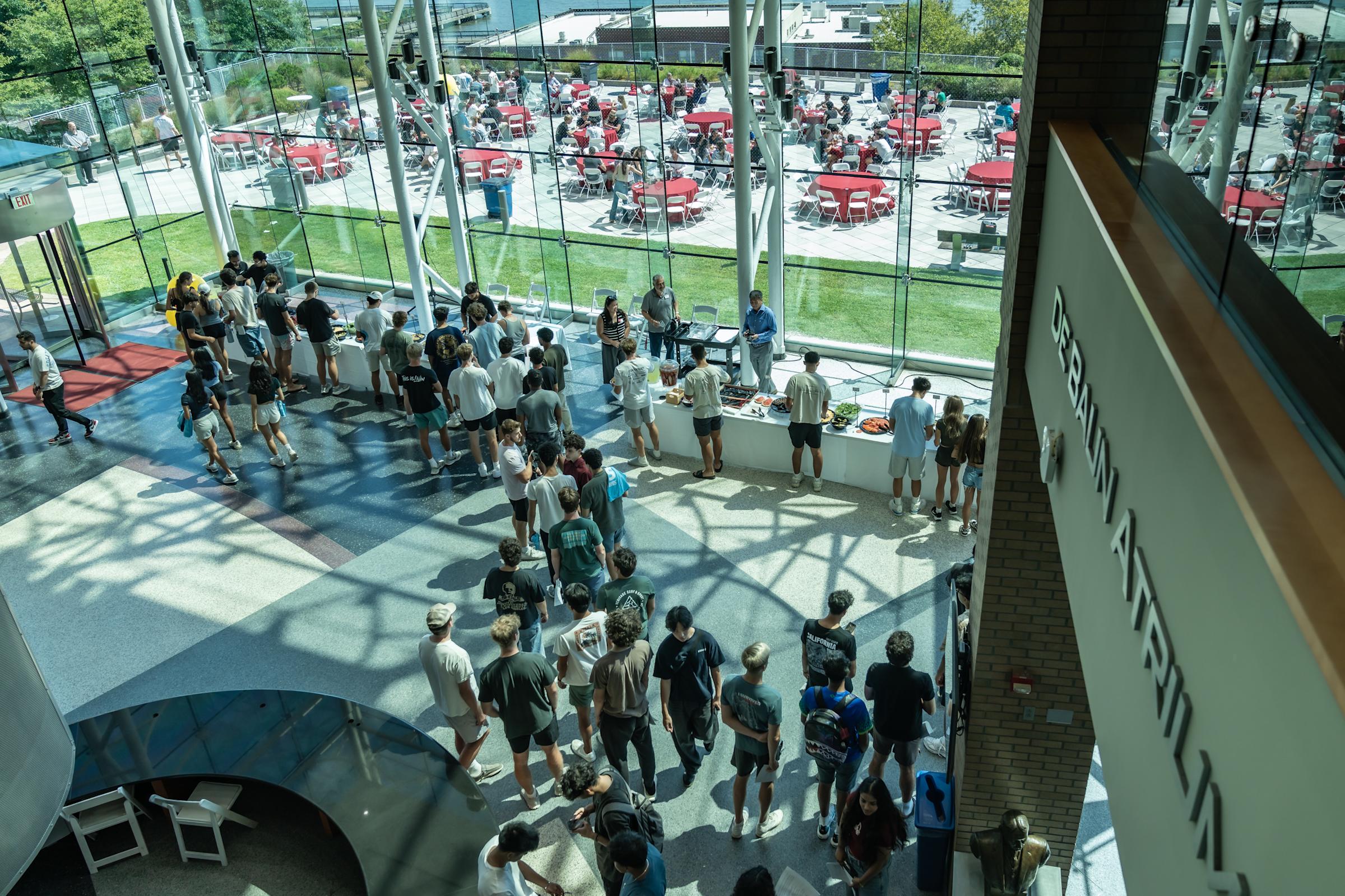 Large group of students queuing in modern glass-walled building lobby, with outdoor dining area featuring red tables visible through windows.