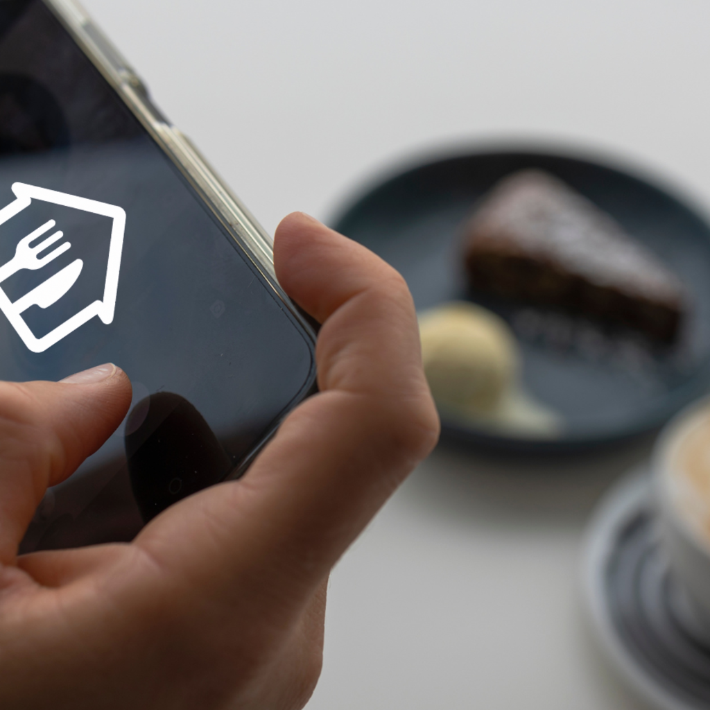 A hand holding up a mobile phone in a restaurant in front of a place setting while a person reads in the background.