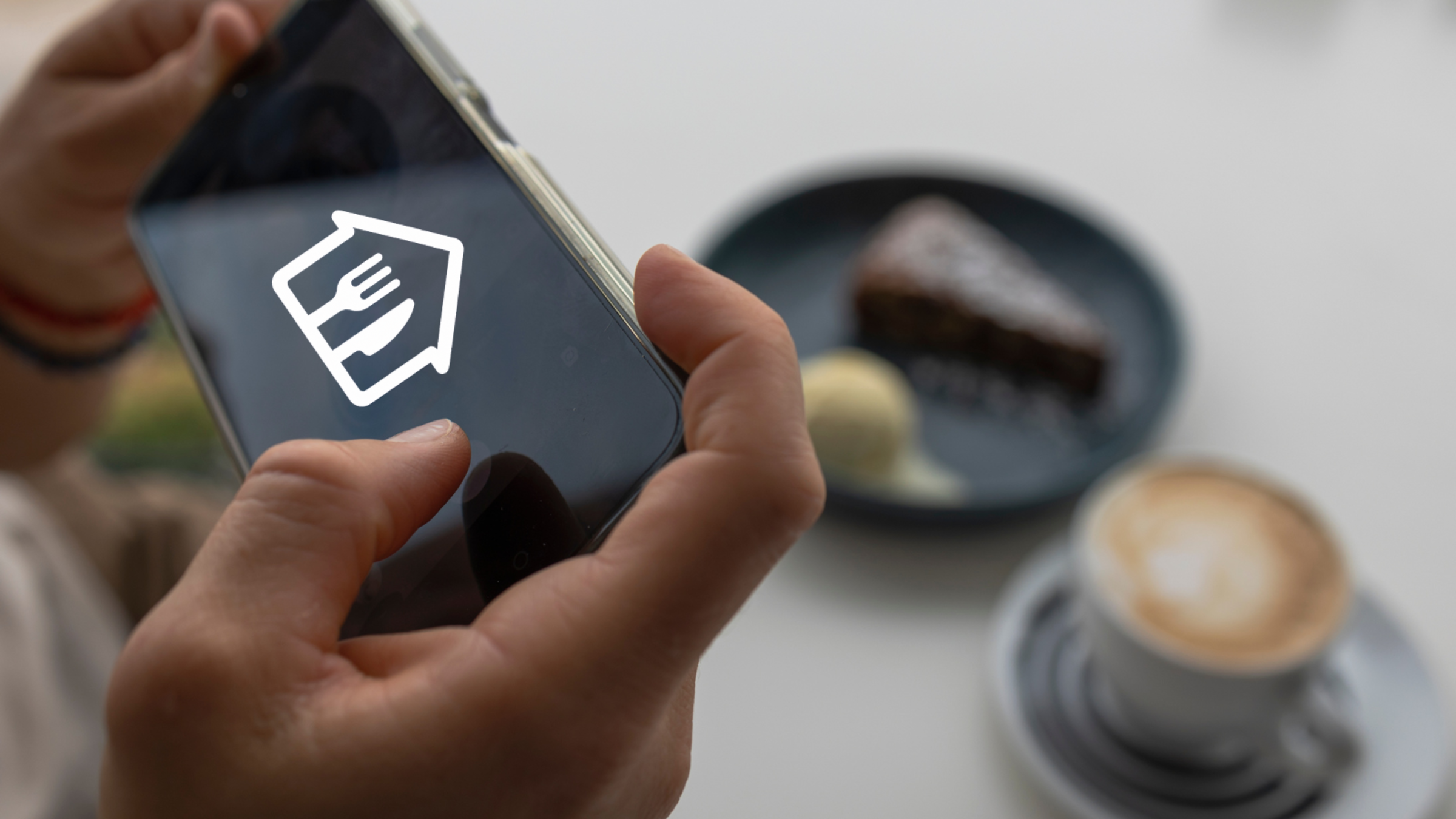 A hand holding up a mobile phone in a restaurant in front of a place setting while a person reads in the background.