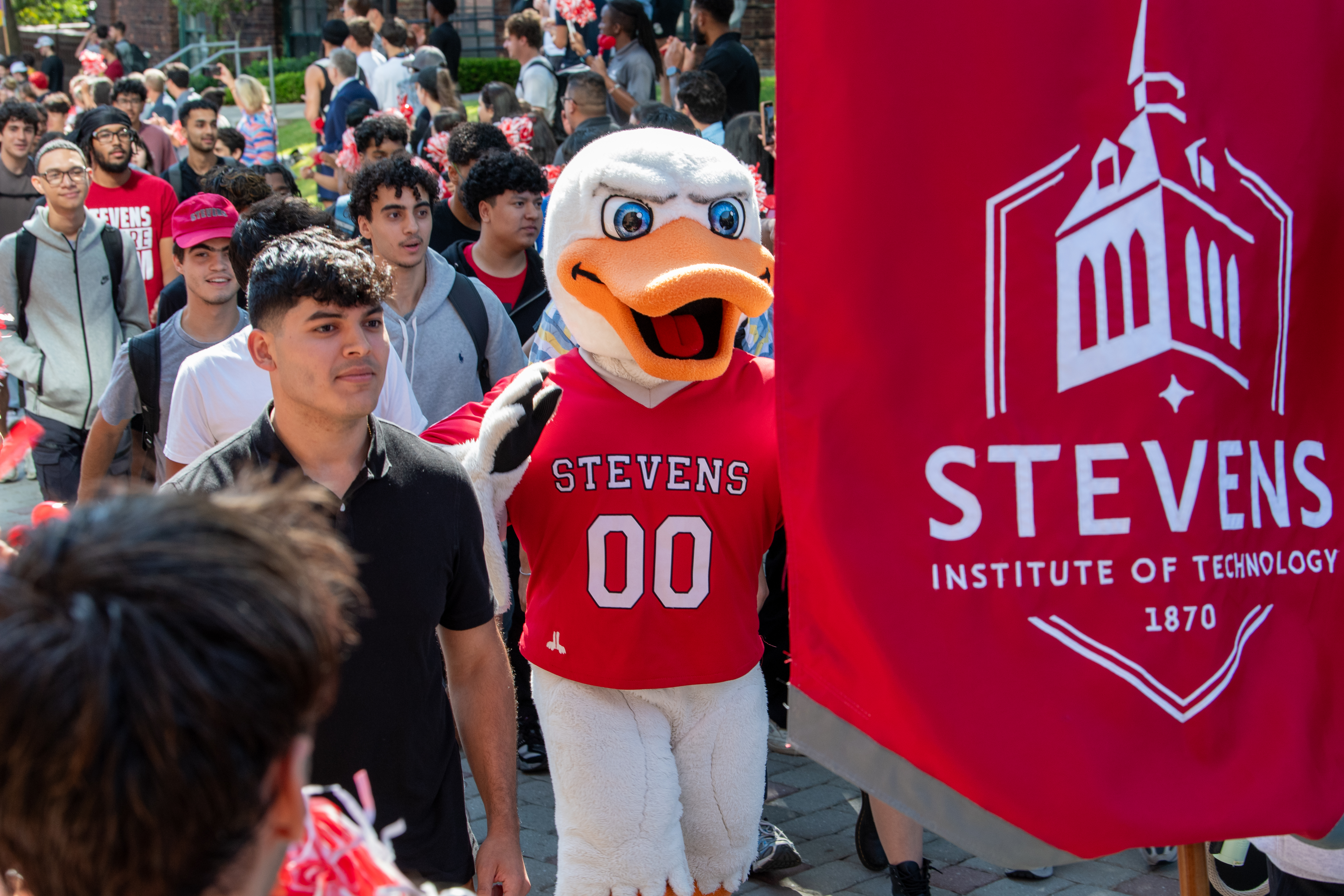 Students walk and cheer with duck mascot in springtime holding banner 