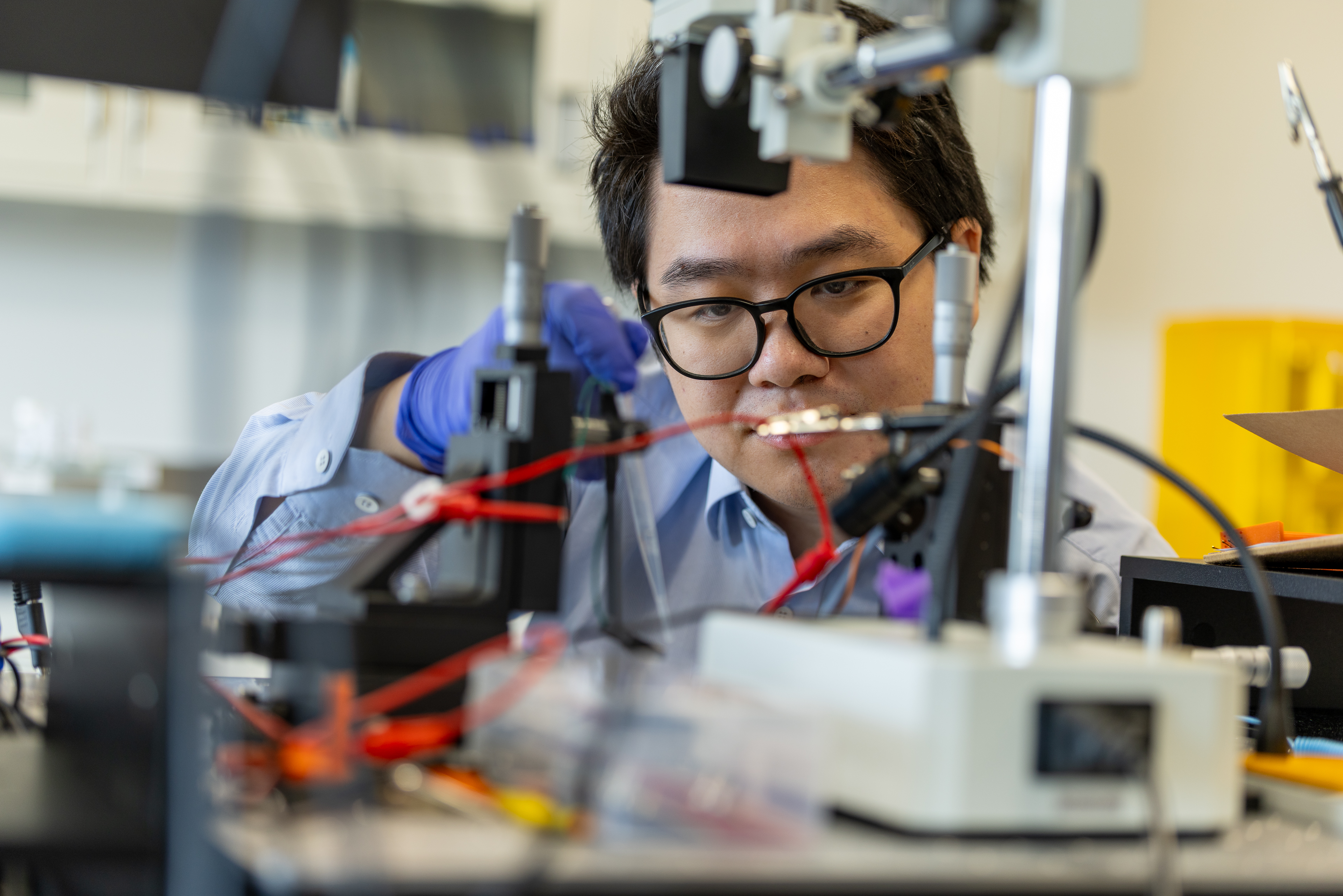 A student wearing glasses holding a pipette in a lab behind a foreground with lab equipment.
