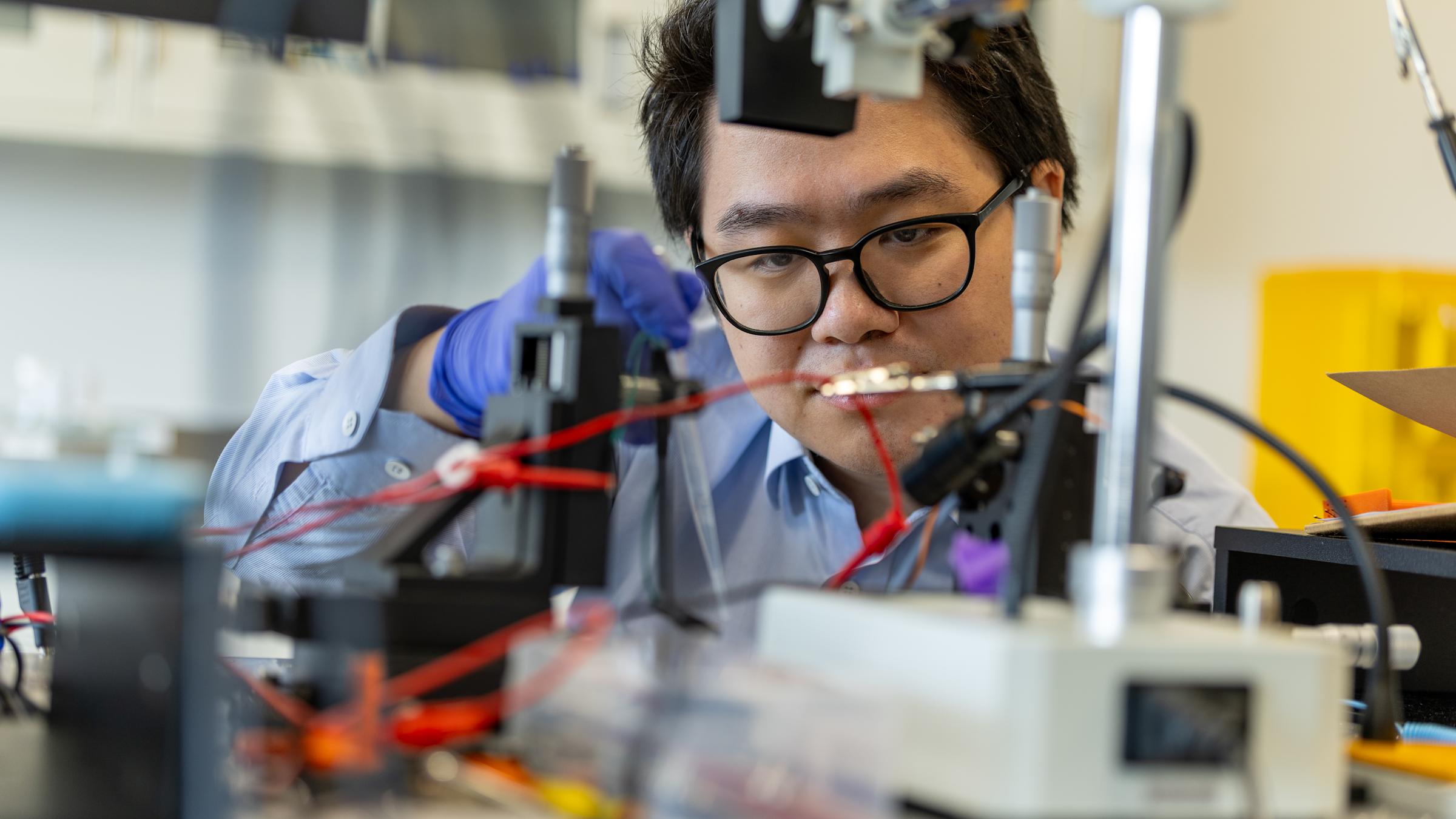 A student wearing glasses holding a pipette in a lab behind a foreground with lab equipment.