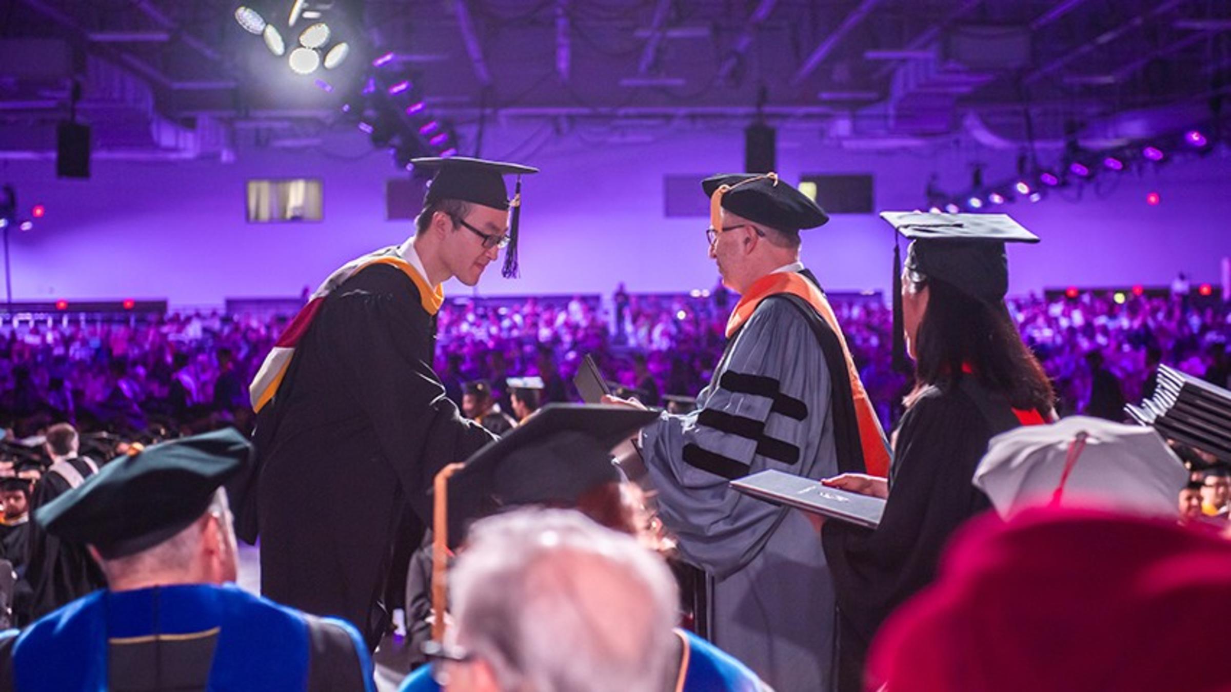 A view from backstage of a diploma being handed to a male student. The audience is visible as a large crowd in the background.
