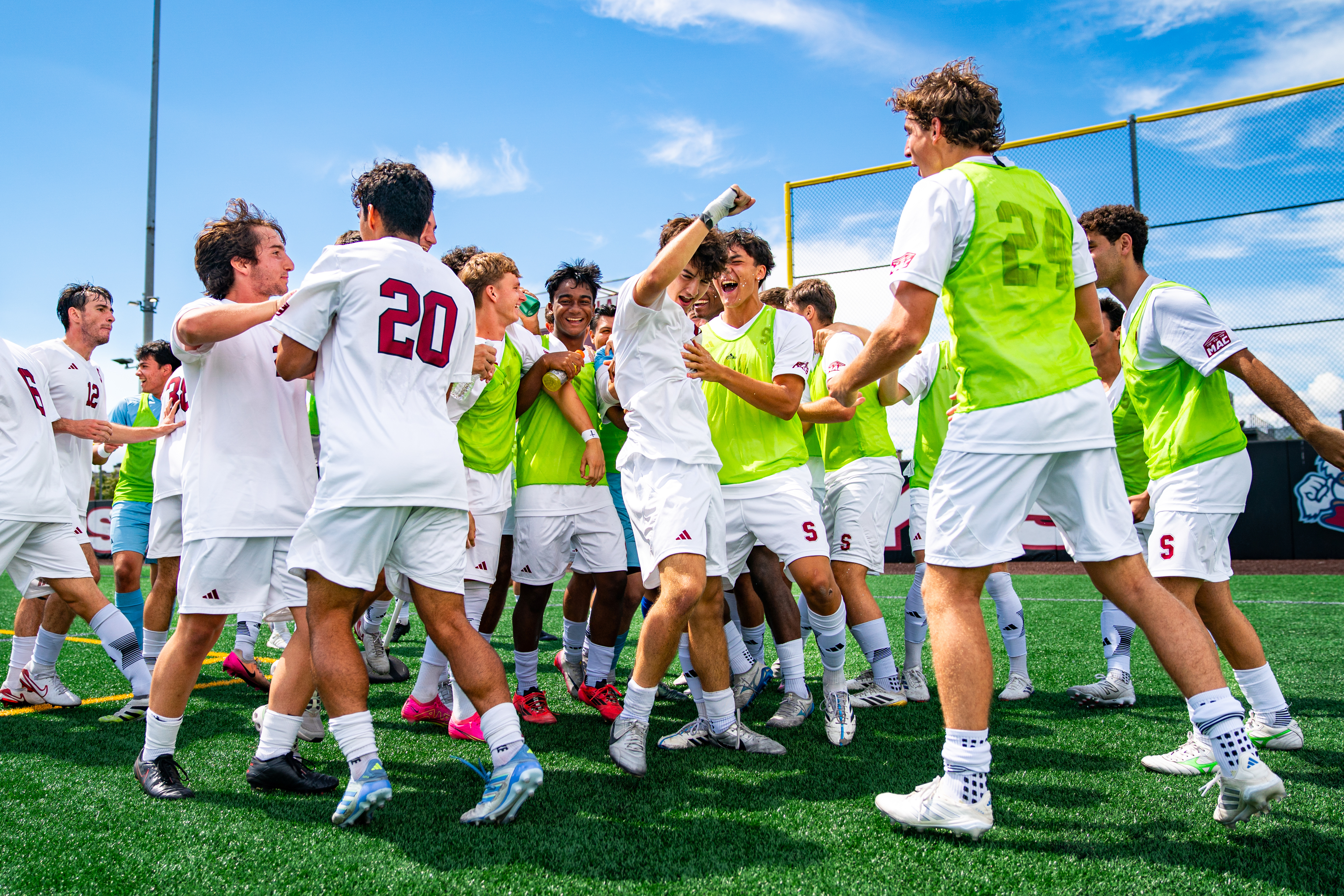 Members of the Stevens men's soccer team gather in a circle on an outdoor turf field, celebrating together. Players wear white uniforms, with several players in bright green pinnies.