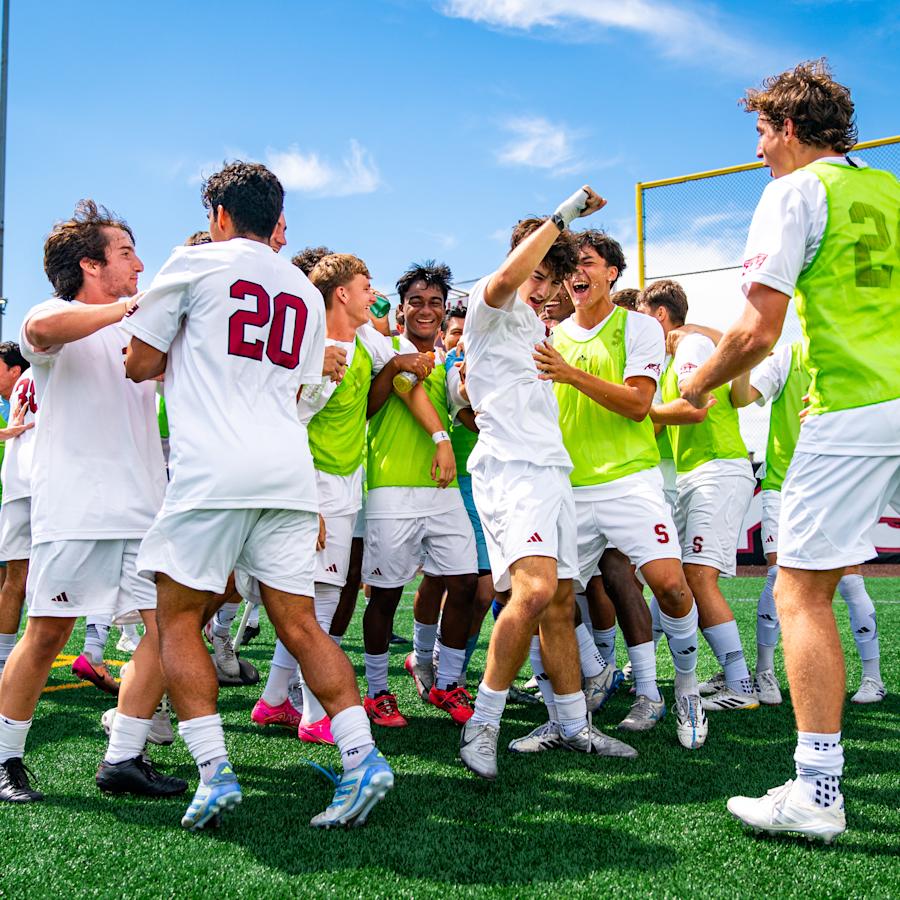Members of the Stevens men's soccer team gather in a circle on an outdoor turf field, celebrating together. Players wear white uniforms, with several players in bright green pinnies.