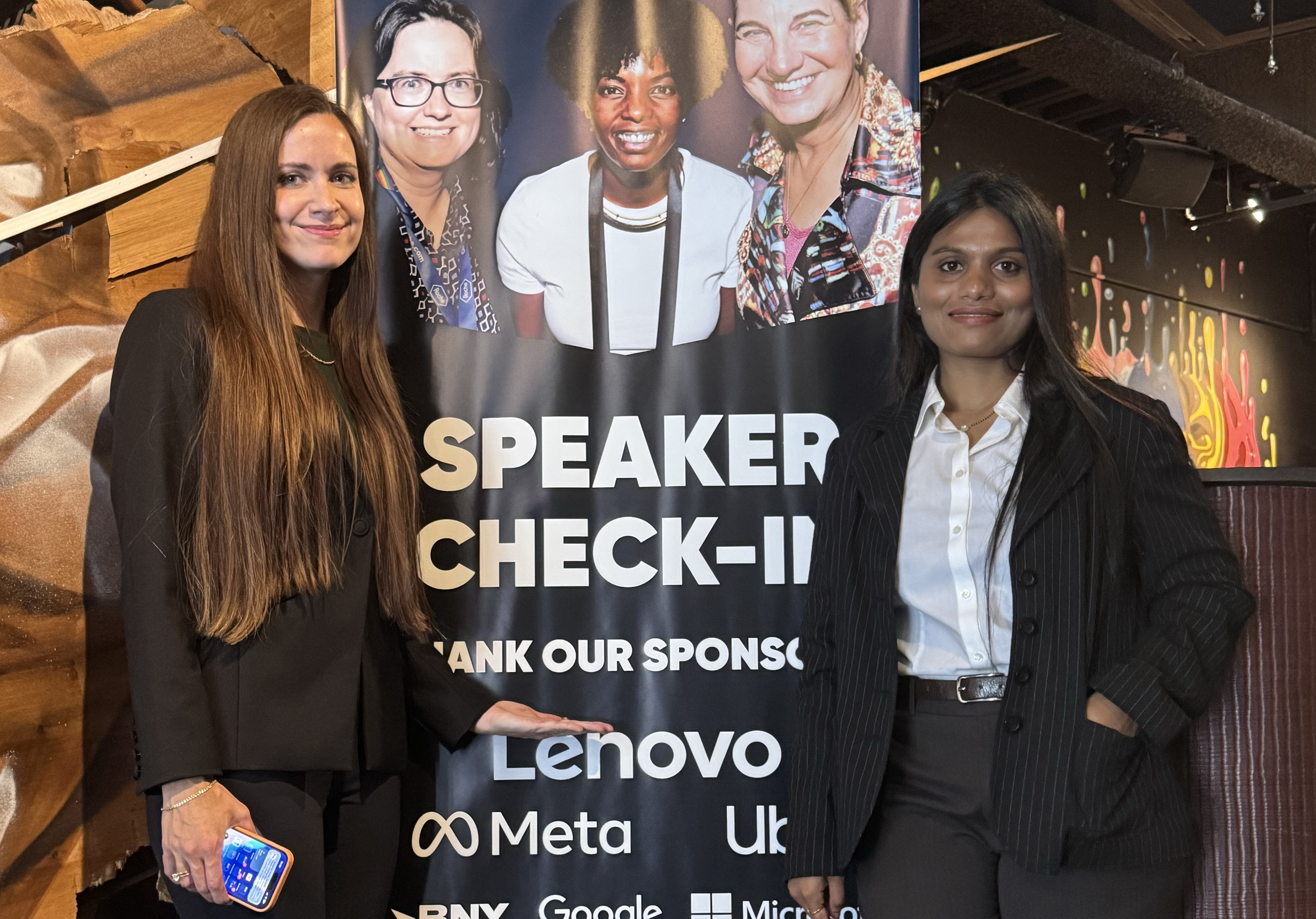 Two women in dark business attire pose beside a "Speaker Check-In" event banner listing sponsors including Lenovo, Meta, Google, and Microsoft. The banner features photos of three women speakers at the top, set in a warmly lit venue with rustic wood decor.