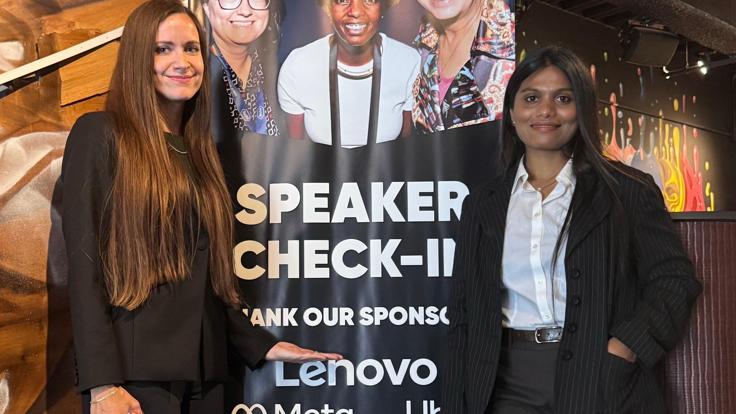 Two women in dark business attire pose beside a "Speaker Check-In" event banner listing sponsors including Lenovo, Meta, Google, and Microsoft. The banner features photos of three women speakers at the top, set in a warmly lit venue with rustic wood decor.