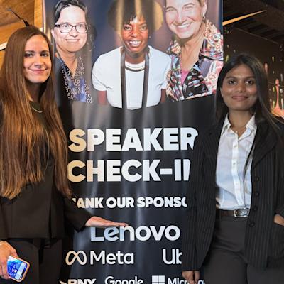 Two women in dark business attire pose beside a "Speaker Check-In" event banner listing sponsors including Lenovo, Meta, Google, and Microsoft. The banner features photos of three women speakers at the top, set in a warmly lit venue with rustic wood decor.