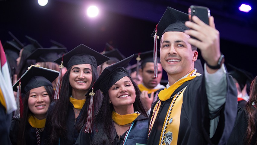 Three female students pose for a selfie taken by a fourth student. All are in their graduation regalia.