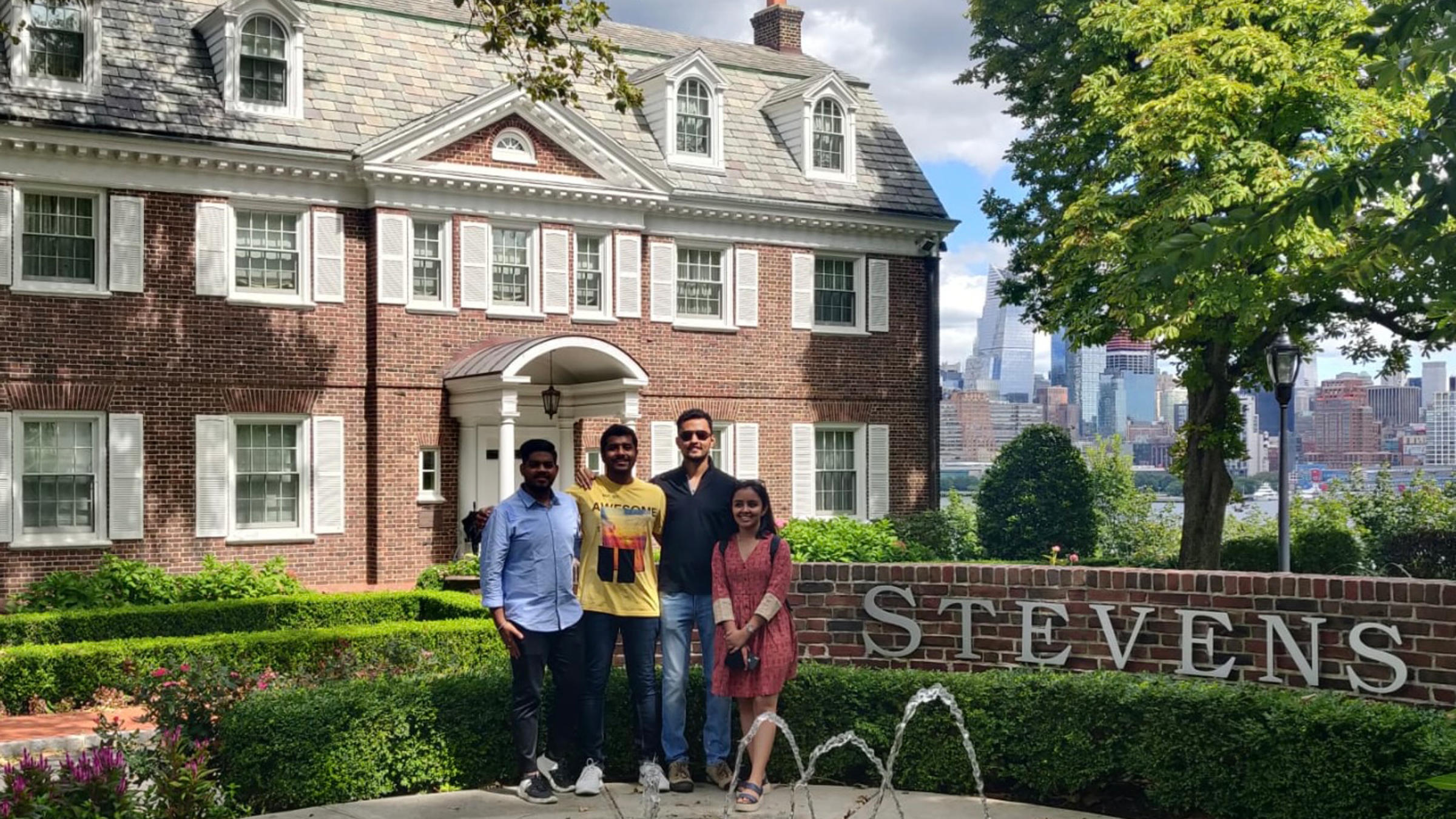 Rhythm Goyal stands with her family in front of the fountain on the Stevens campus with the New York City skyline visible in the background.