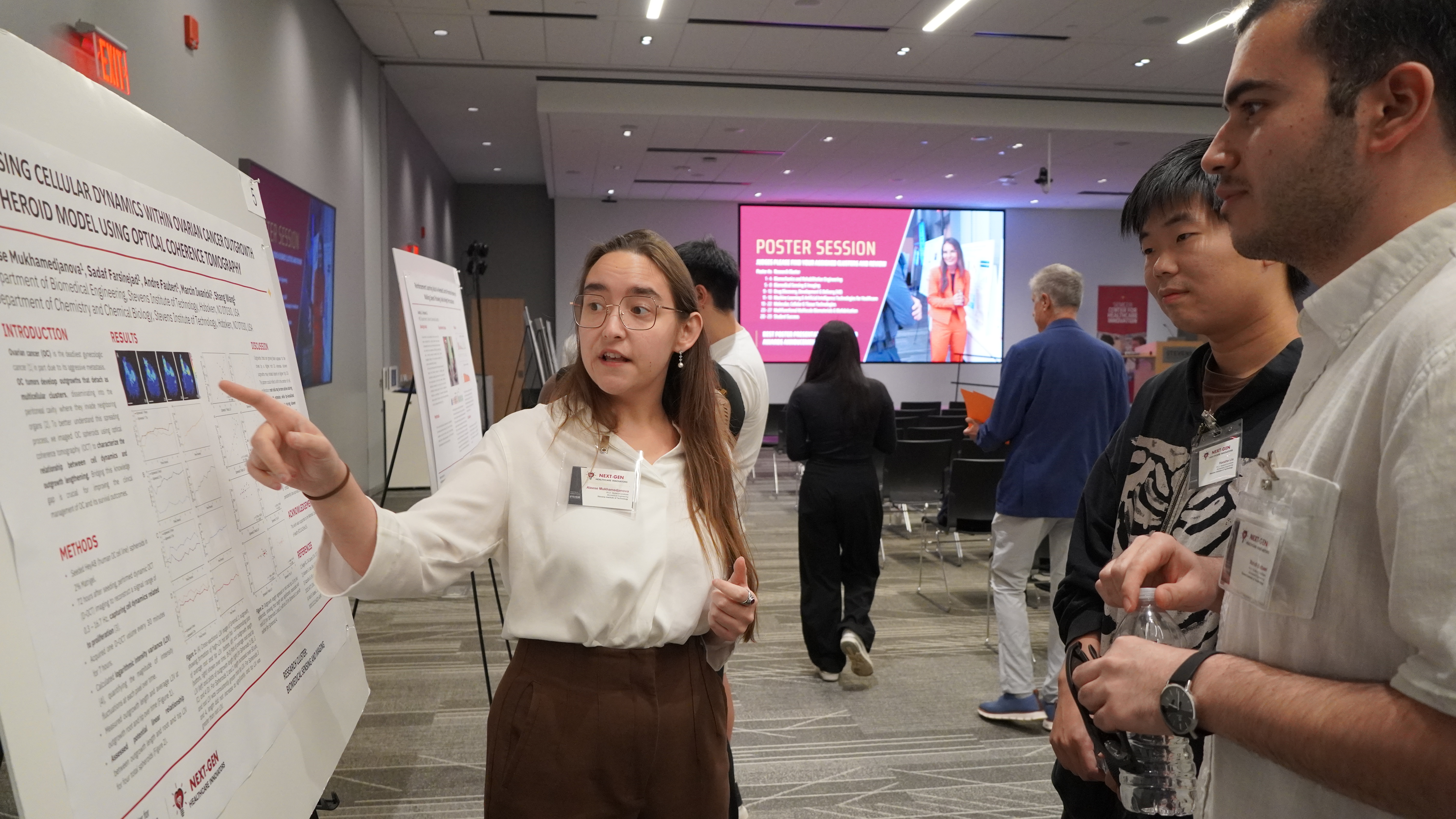 A student with glasses explains their poster presentation to other observers.