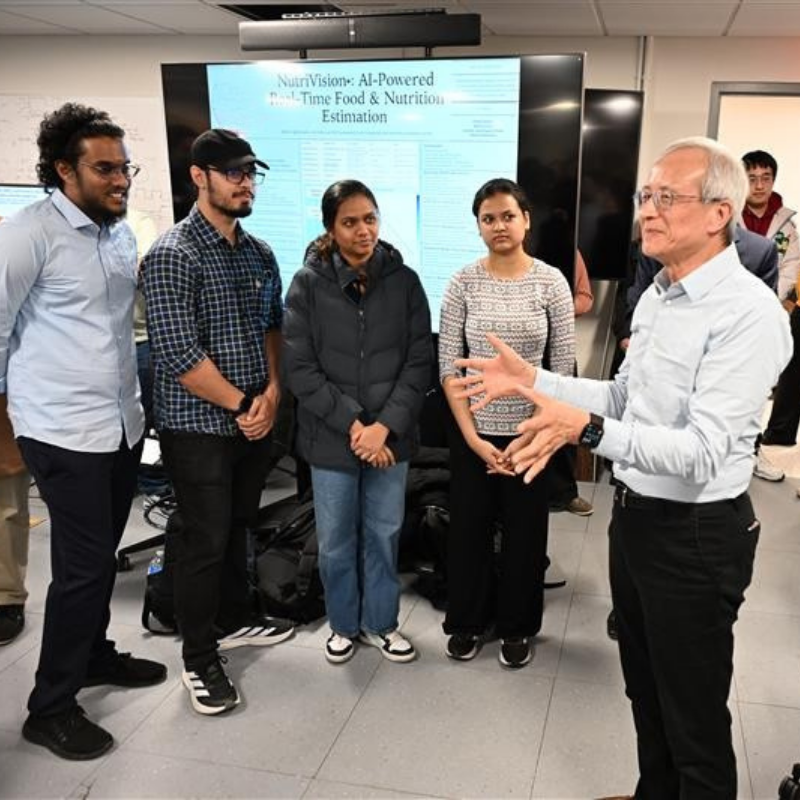 A group of students on the left listening to a professor speak on the right, who is gesturing with his hands and another professor is standing behind him.