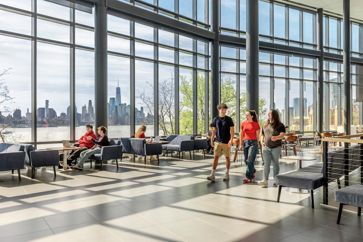Students walk inside the University Center Complex on a sunny day, the NYC skyline in the background.