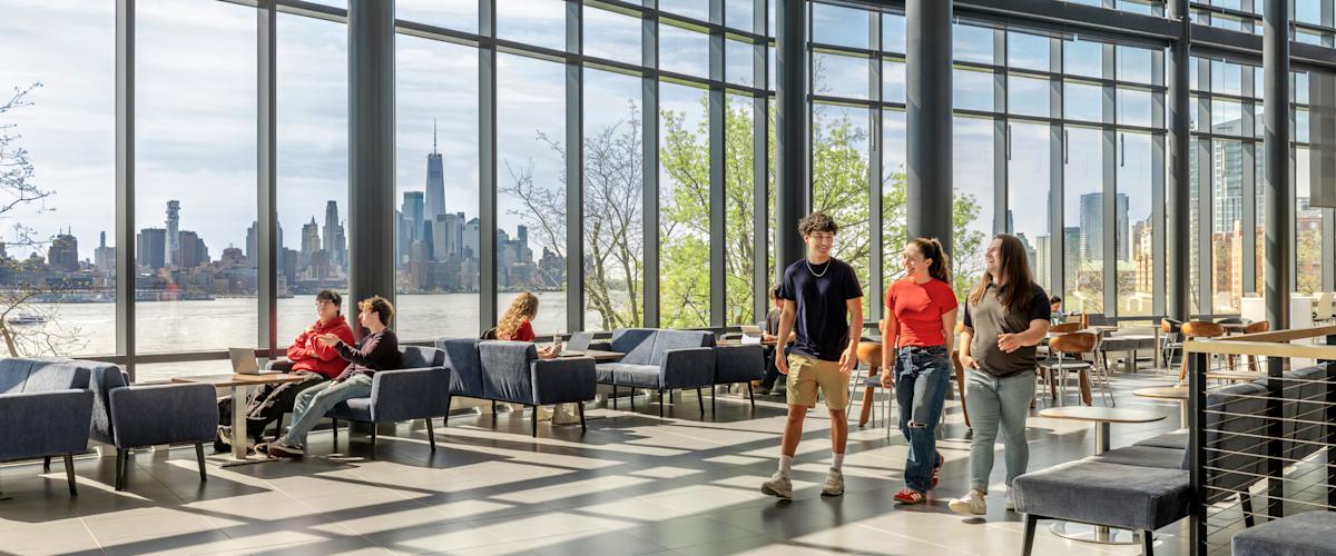 Students walk inside the University Center Complex on a sunny day, the NYC skyline in the background.