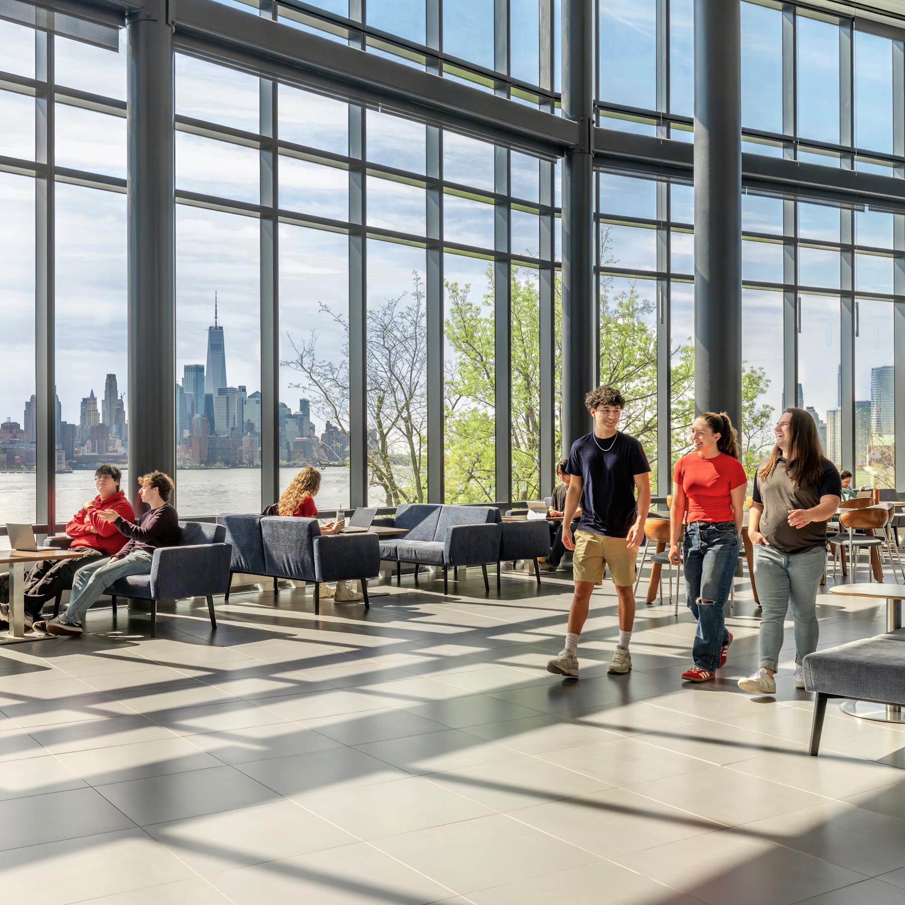 Students walk inside the University Center Complex on a sunny day, the NYC skyline in the background.