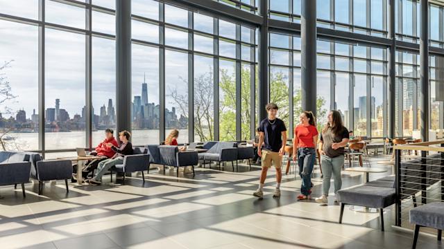Students walk inside the University Center Complex on a sunny day, the NYC skyline in the background.