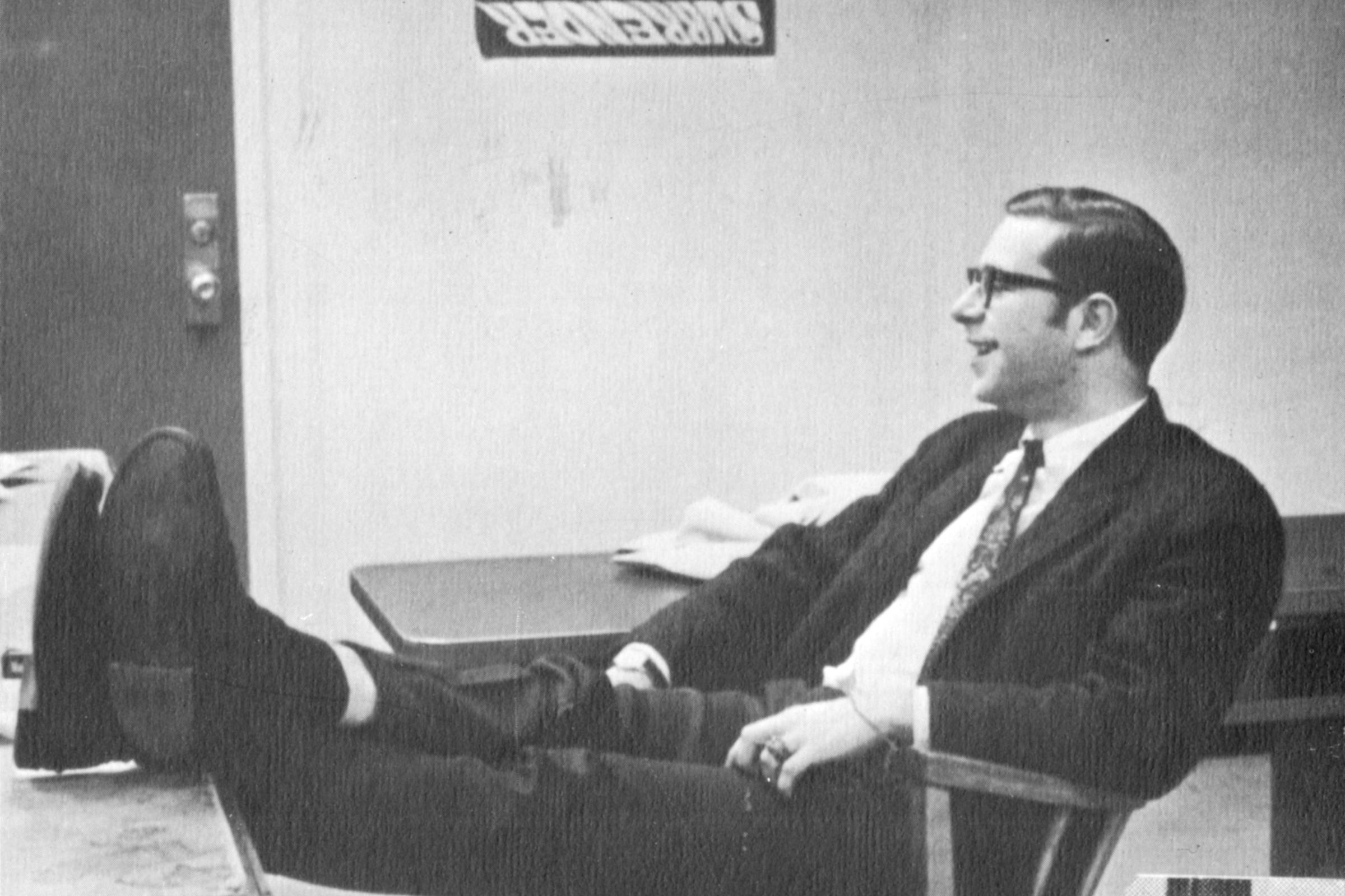 Man wearing glasses sits in chair with feet up on table.