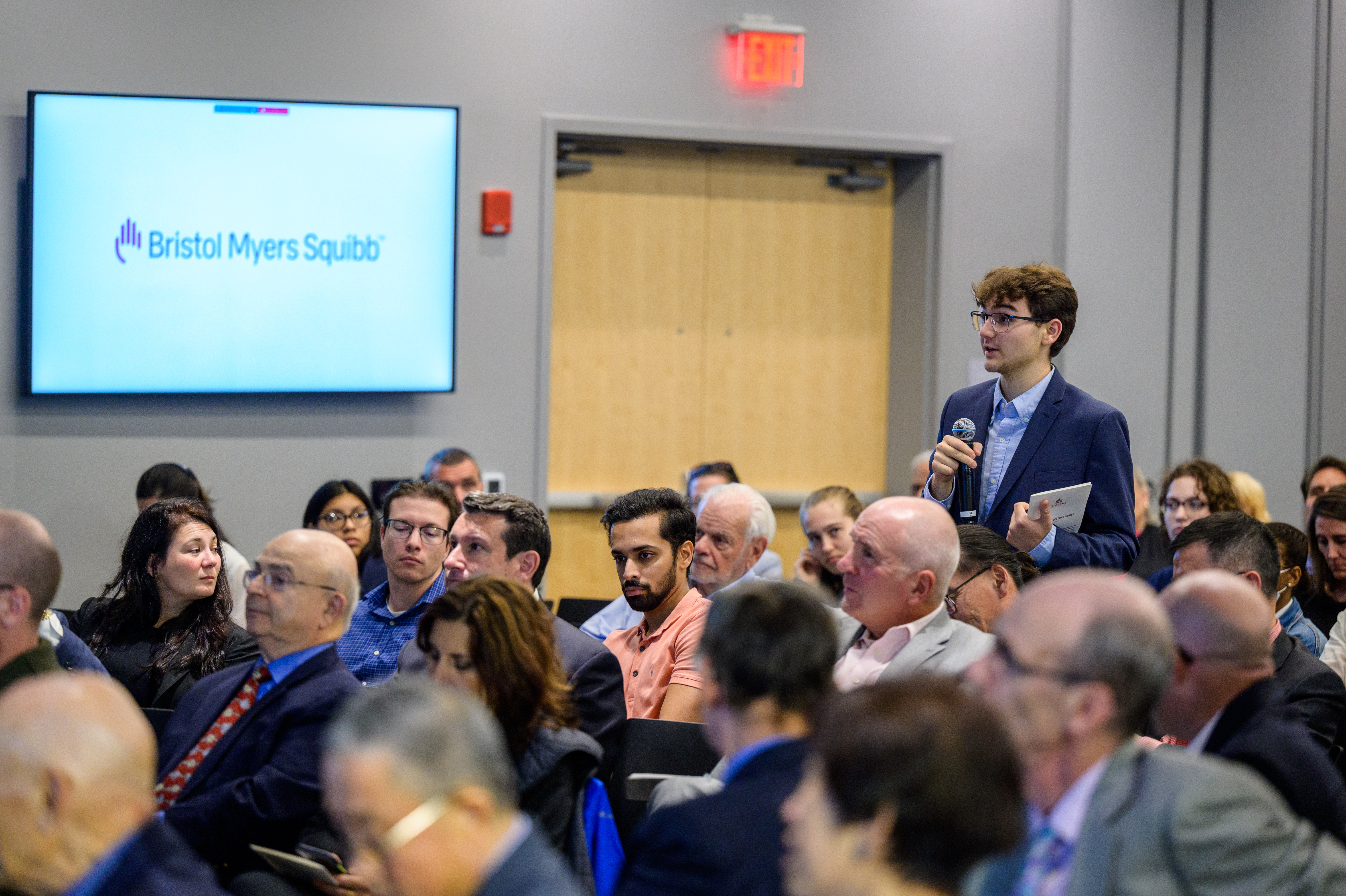 A man in a suit speaks into a microphone during a conference, with a Bristol Myers Squibb logo visible in the background.
