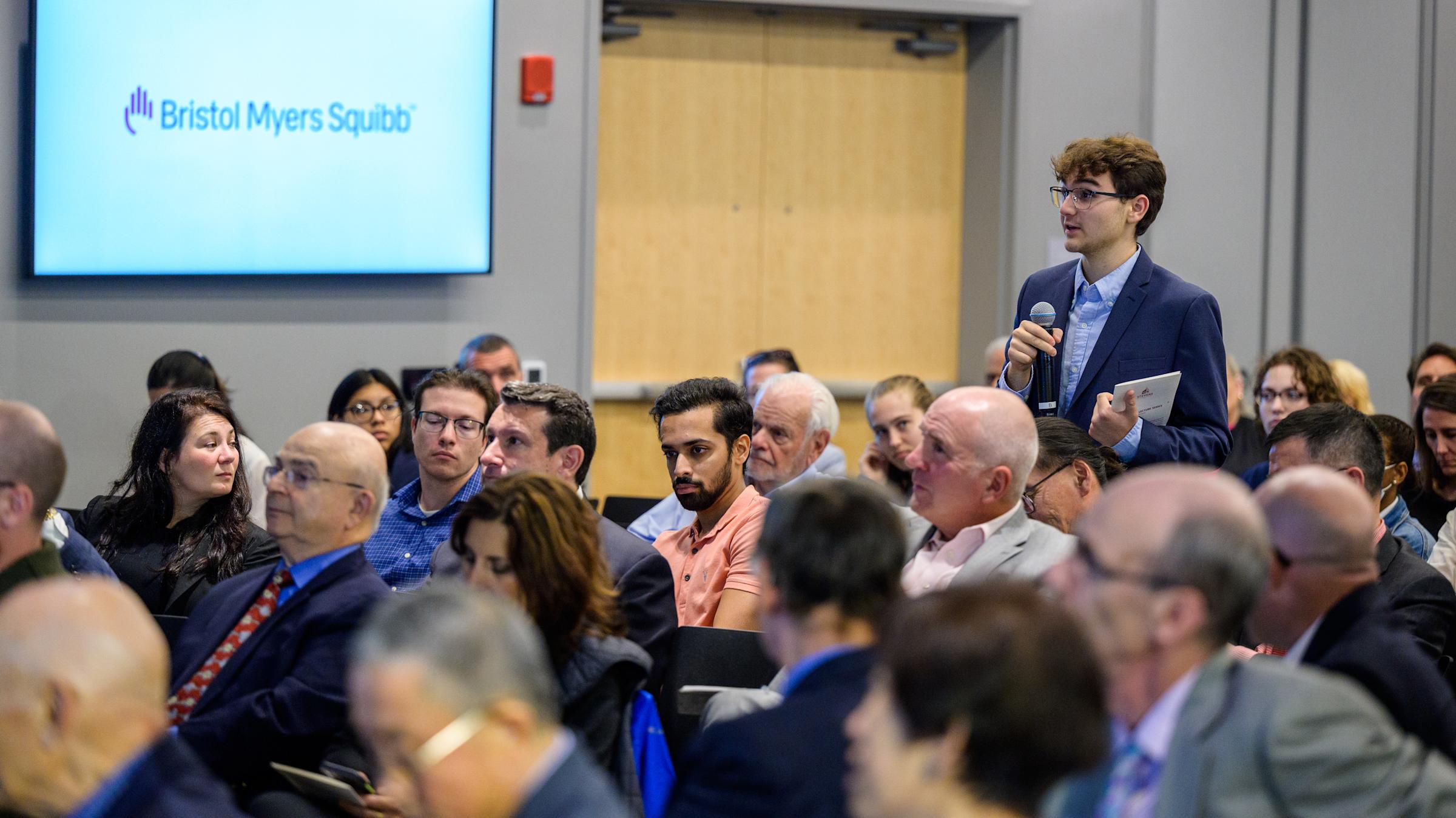 A man in a suit speaks into a microphone during a conference, with a Bristol Myers Squibb logo visible in the background.