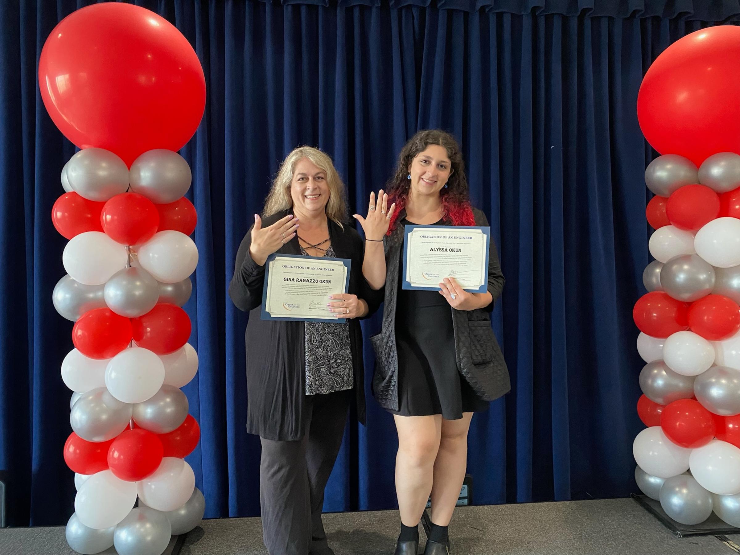 Okun daughter and mother stand together, holding up their certificates, as well as their hands to display their matching rings