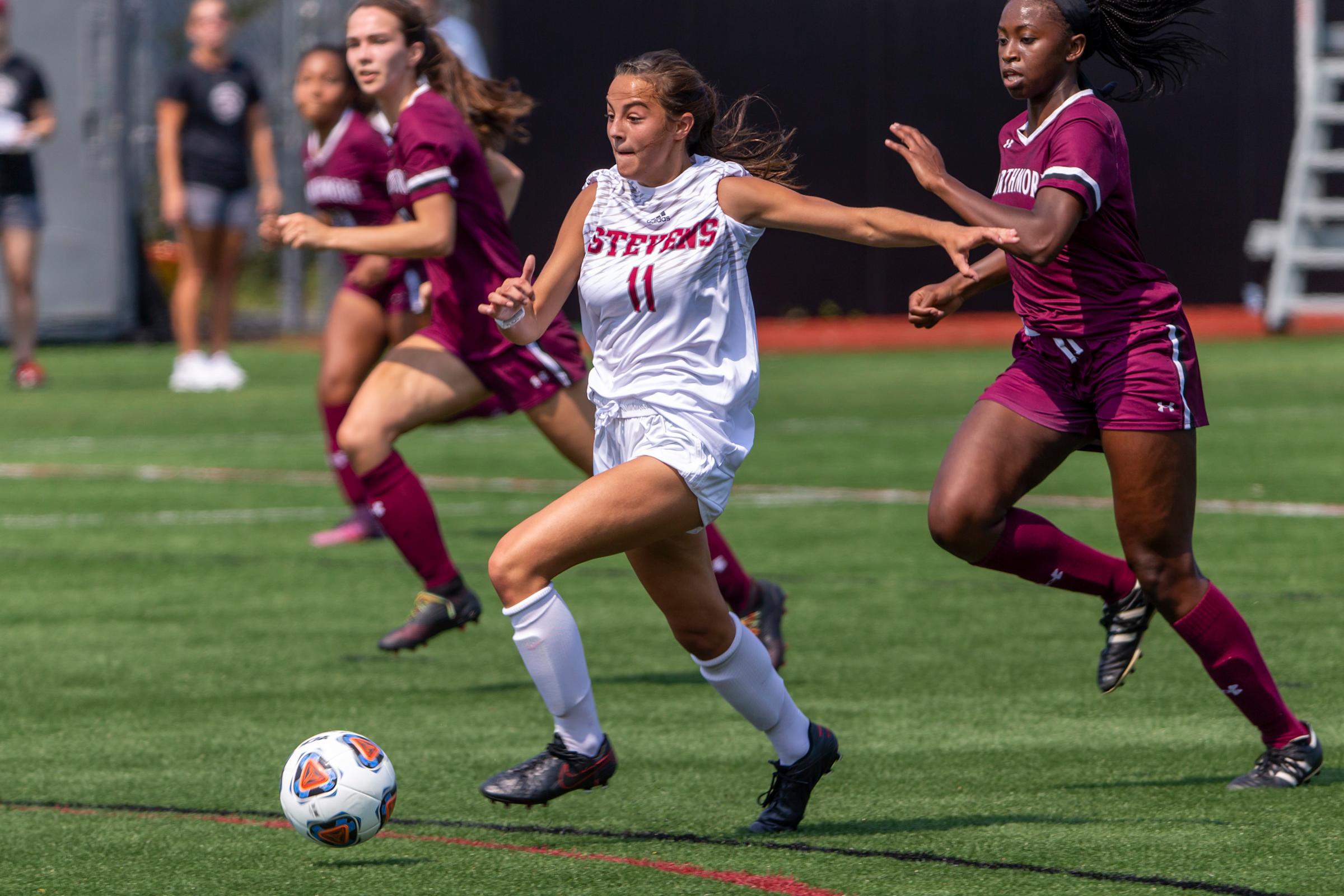 Jenna Mucci jumps on soccer ball, leading over a competing player who is kicking from behind