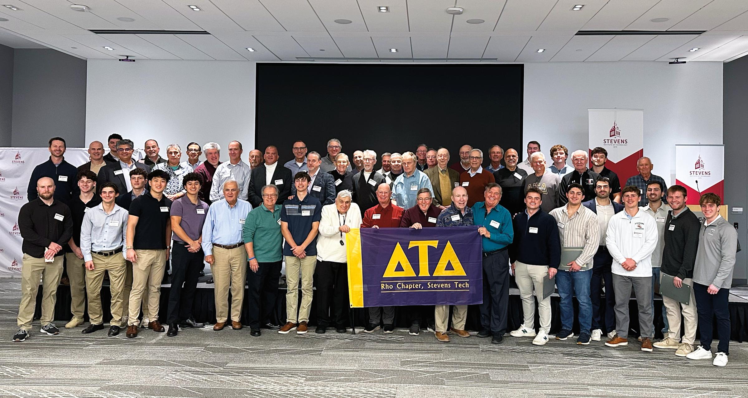 Photo of dozens of Delta Tau Delta fraternity alumni and student members gathered inside the Tech Flex auditorium of the University Center Complex. Several display a purple and gold banner with Delta Tau Delta Greek letters.