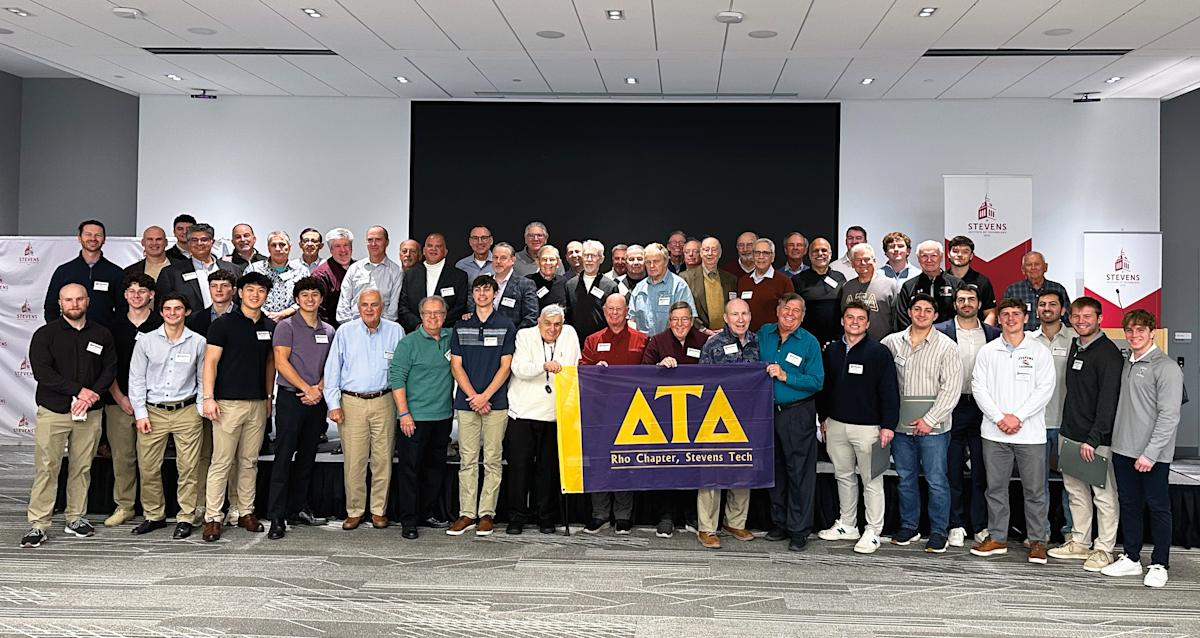 Photo of dozens of Delta Tau Delta fraternity alumni and student members gathered inside the Tech Flex auditorium of the University Center Complex. Several display a purple and gold banner with Delta Tau Delta Greek letters.