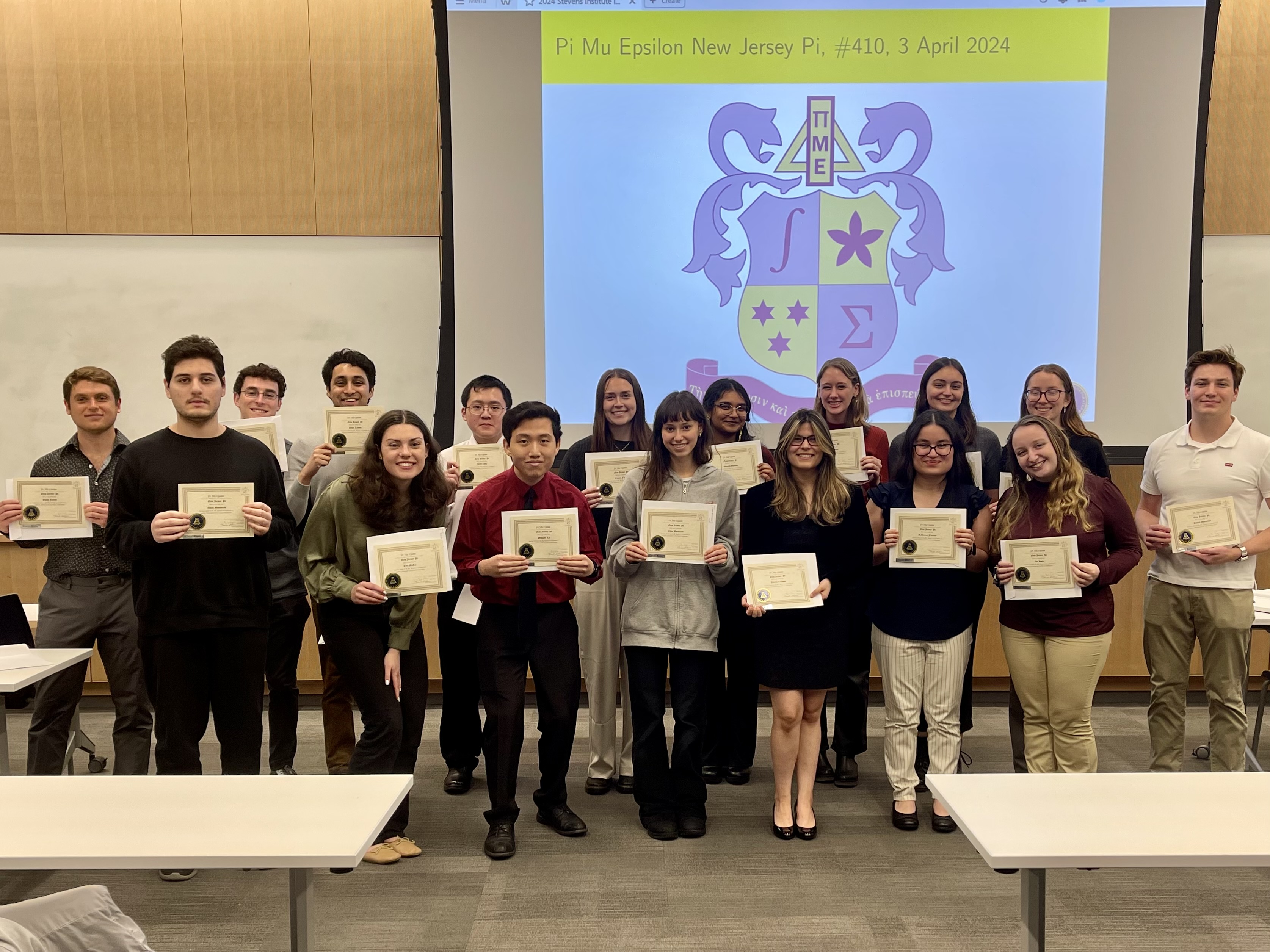 A group of students stand together holding up certificates. They are standing in front of a projector that is displaying a blue and yellow coat of arms.