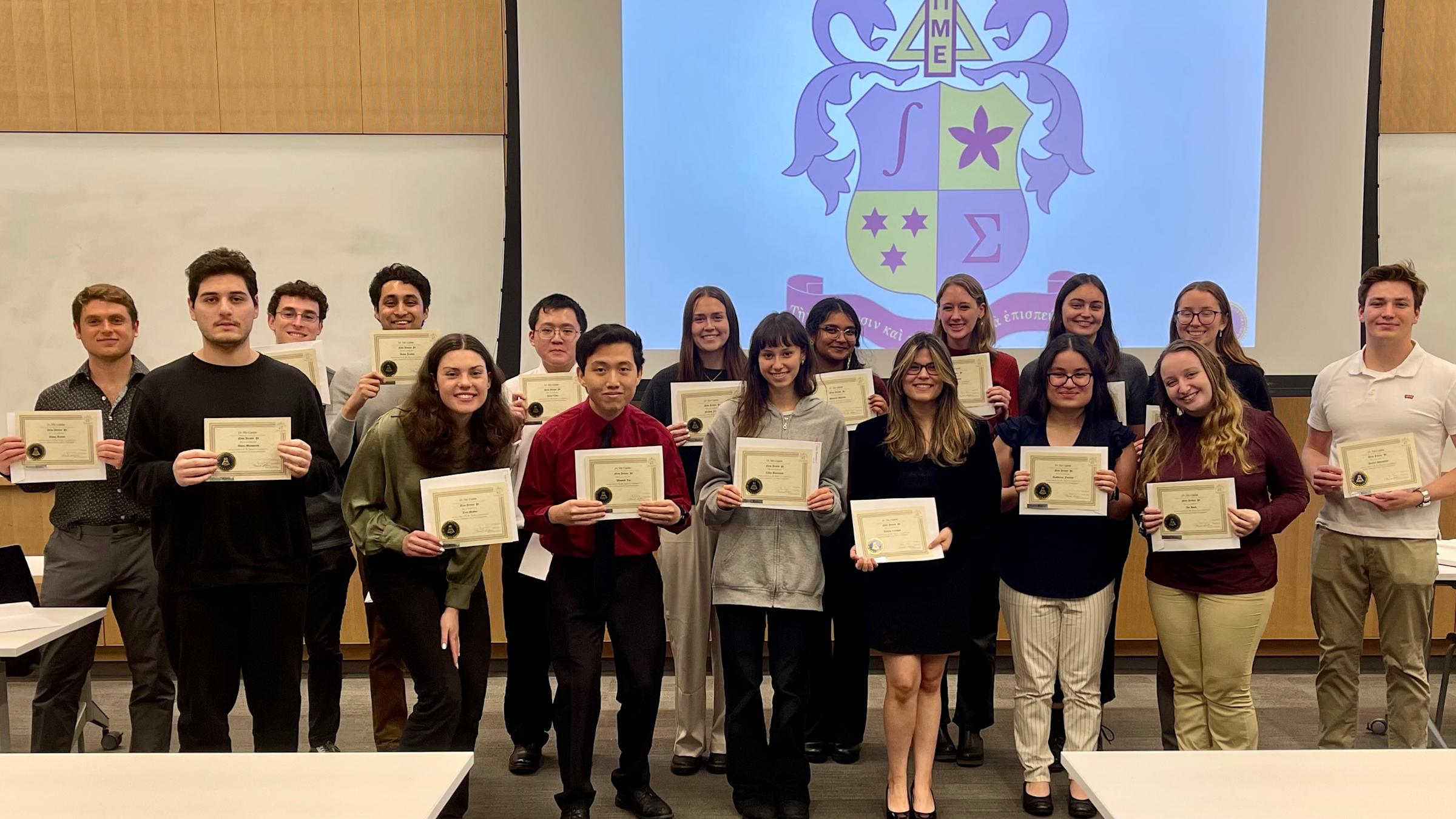 A group of students stand together holding up certificates. They are standing in front of a projector that is displaying a blue and yellow coat of arms.