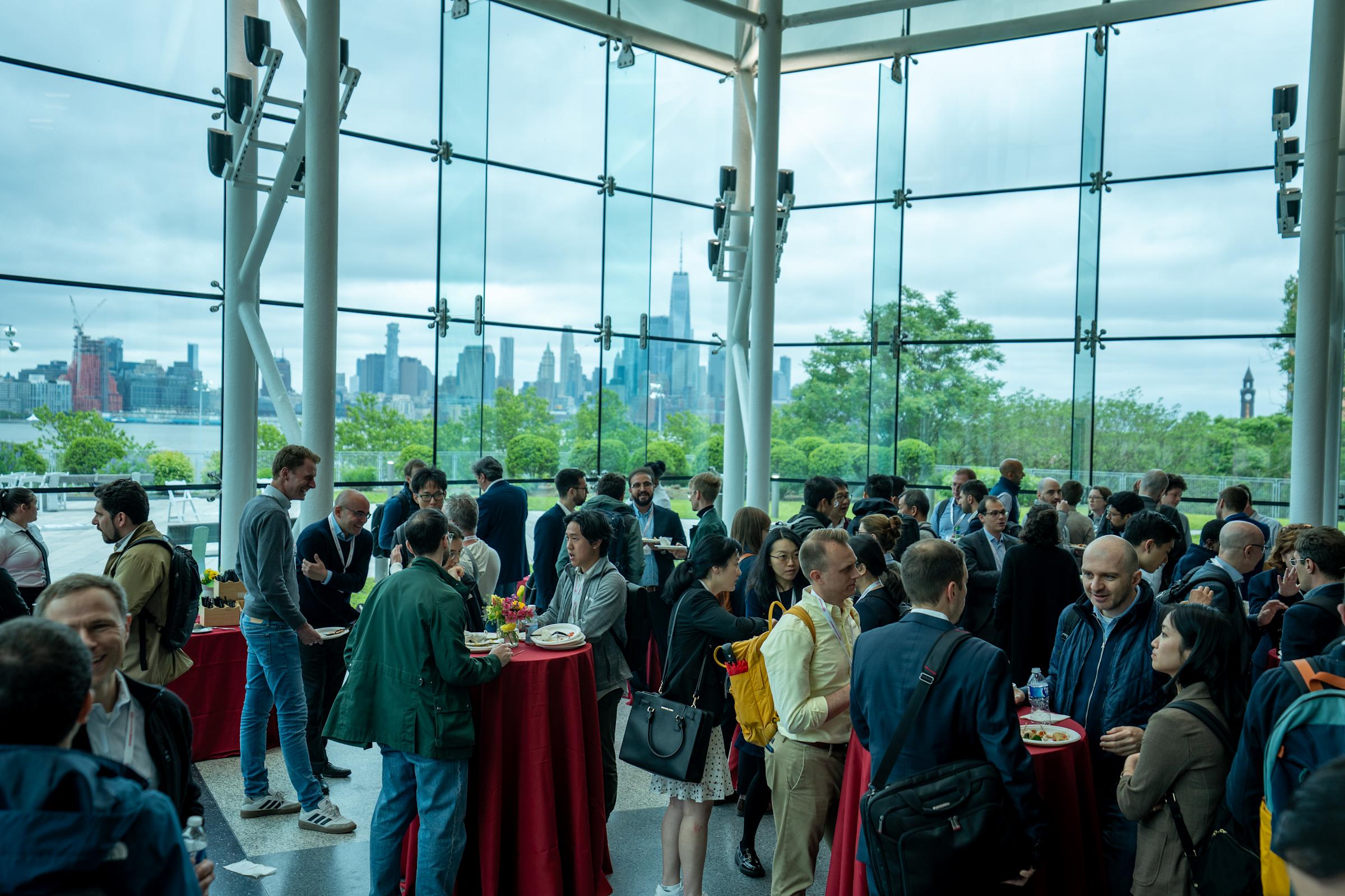 Conference networking reception in a glass-walled venue with Manhattan skyline views. Attendees mingle around red-clothed cocktail tables, eating and socializing in the bright modern space overlooking the city and greenery.