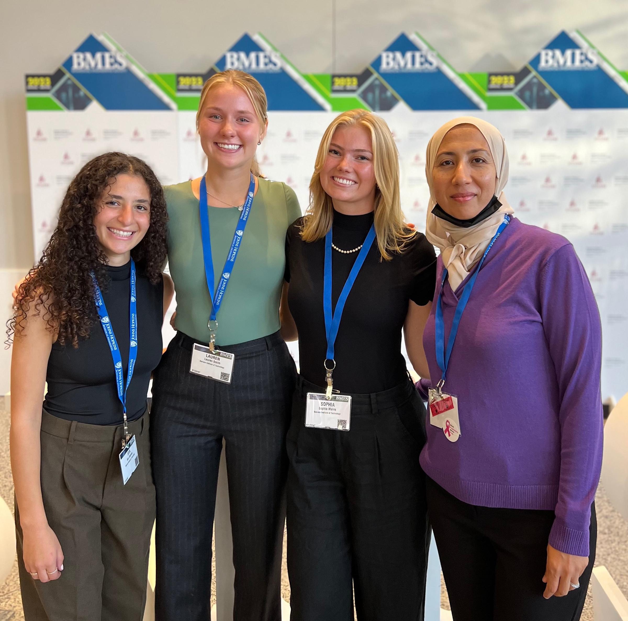 A group of four women stand together in front of a conference poster with "BMES" signage