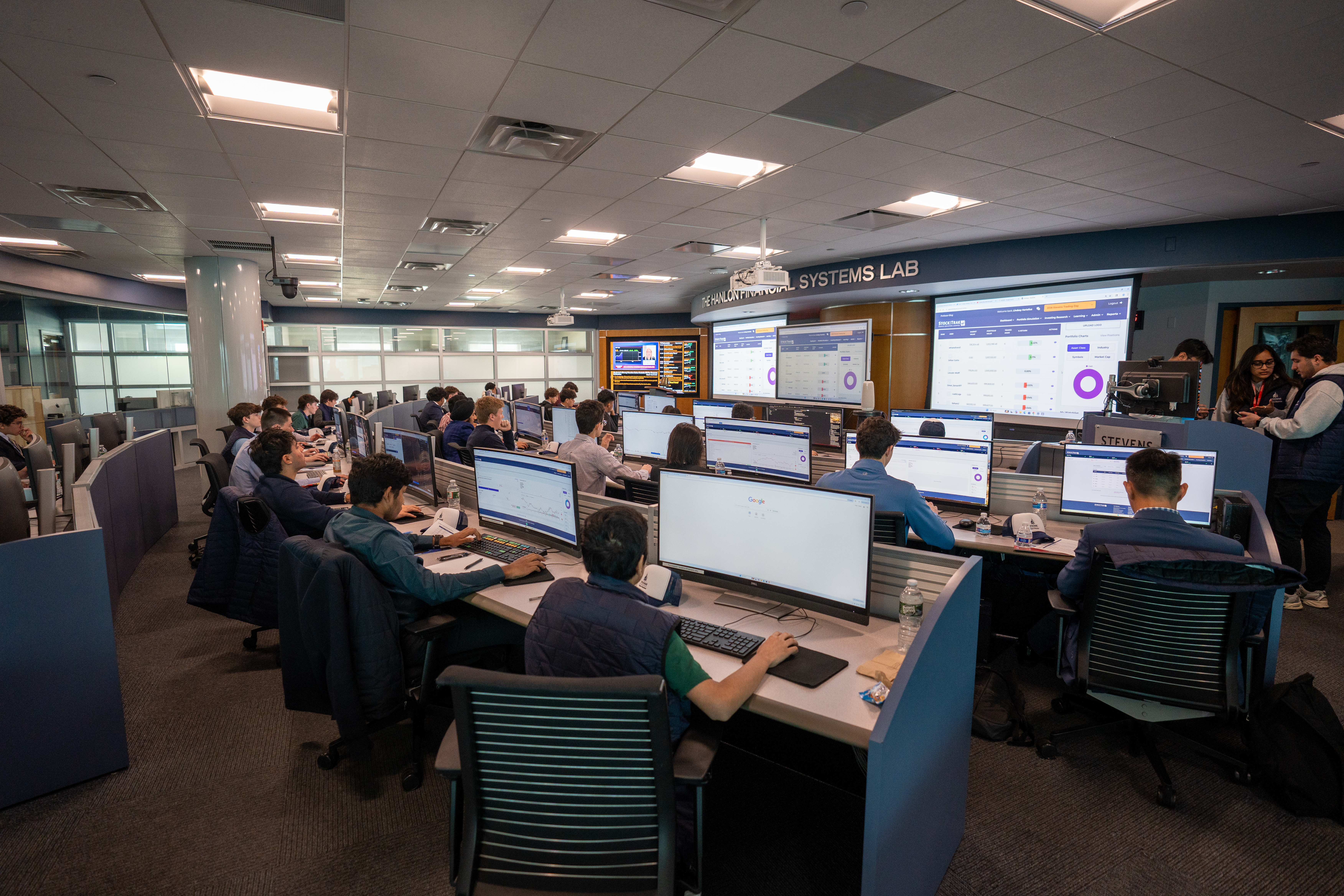 Dozens of students work at tiered trading desks in The Hanlon Financial Systems Lab, each at dual monitors displaying financial data and trading platforms. Large wall-mounted screens show market information and analytics. 