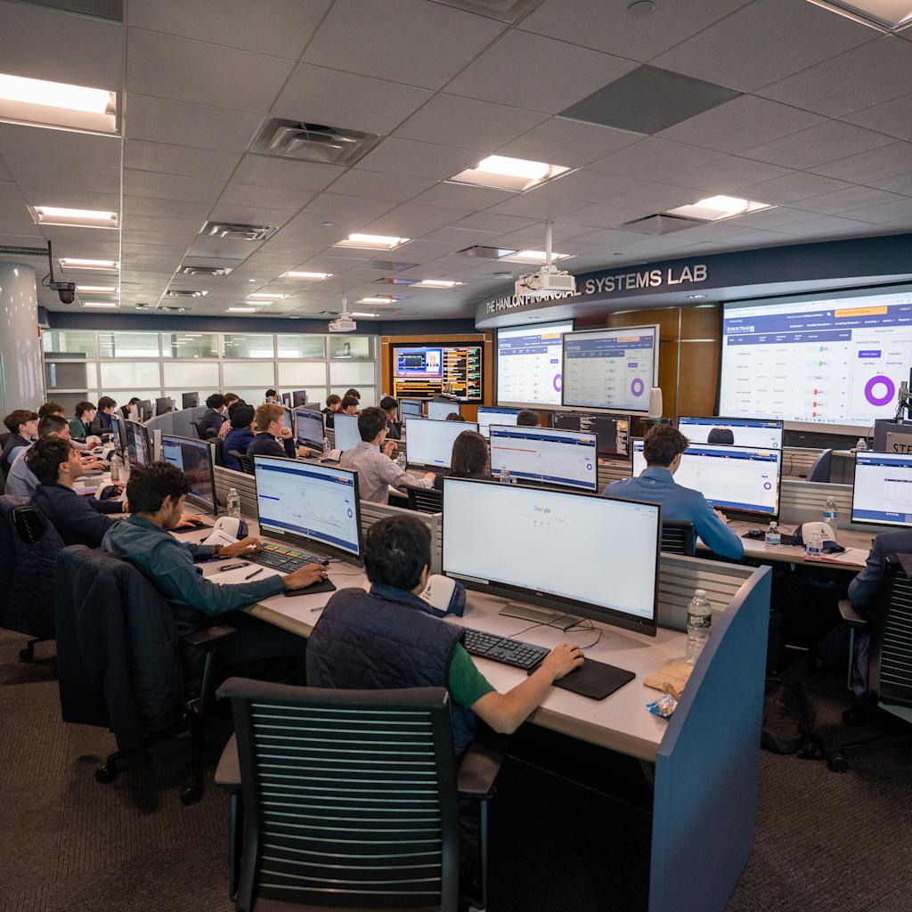Dozens of students work at tiered trading desks in The Hanlon Financial Systems Lab, each at dual monitors displaying financial data and trading platforms. Large wall-mounted screens show market information and analytics.