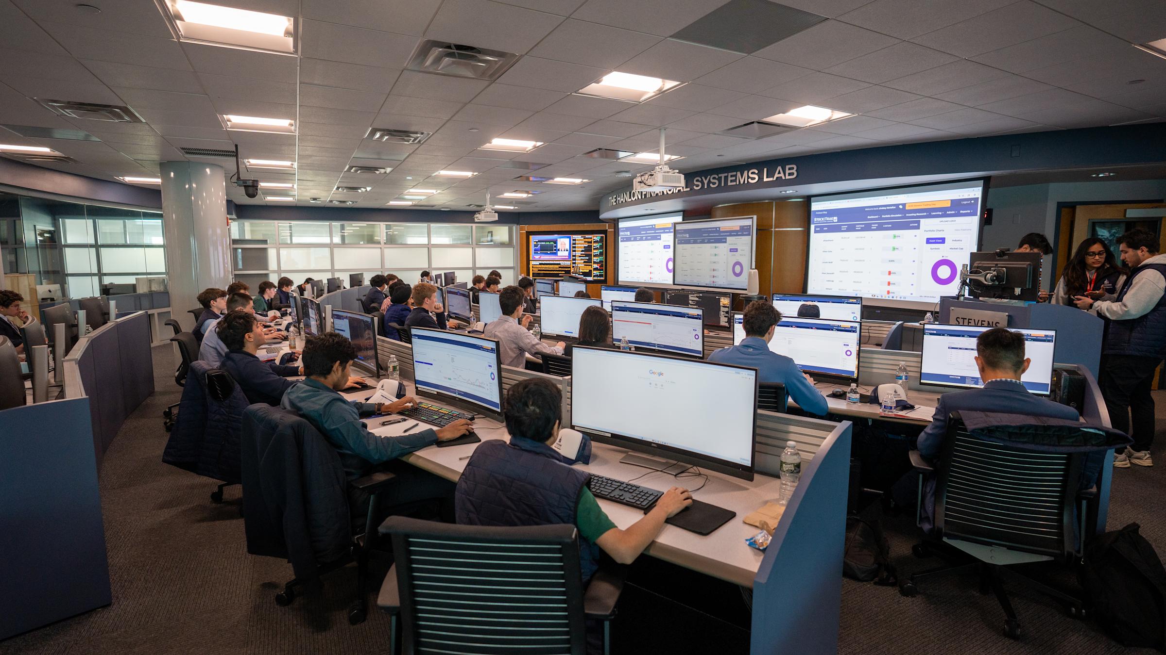 Dozens of students work at tiered trading desks in The Hanlon Financial Systems Lab, each at dual monitors displaying financial data and trading platforms. Large wall-mounted screens show market information and analytics.