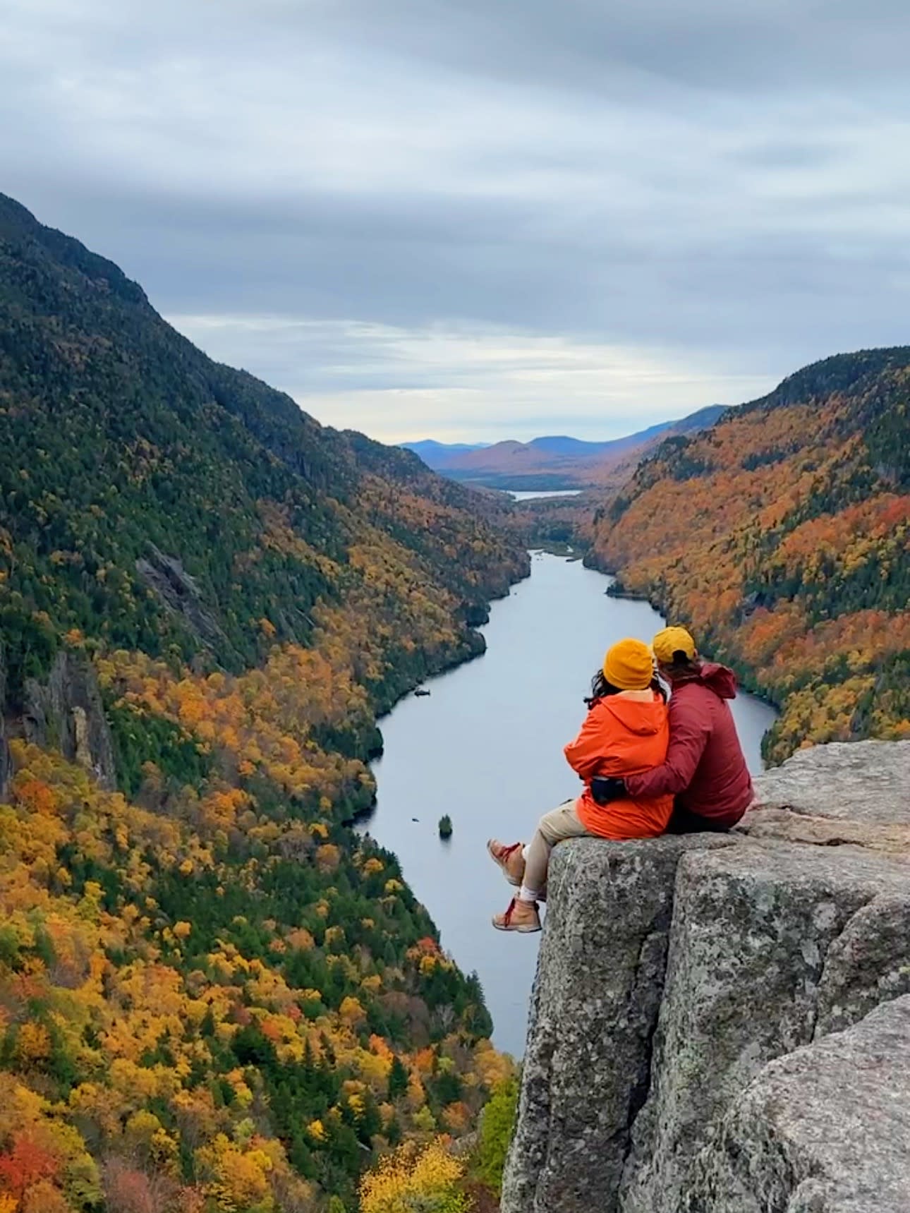 Two hikers in orange and red jackets sit together on a rocky cliff edge, embracing as they overlook a stunning river valley flanked by vibrant autumn foliage in orange, yellow, and green.