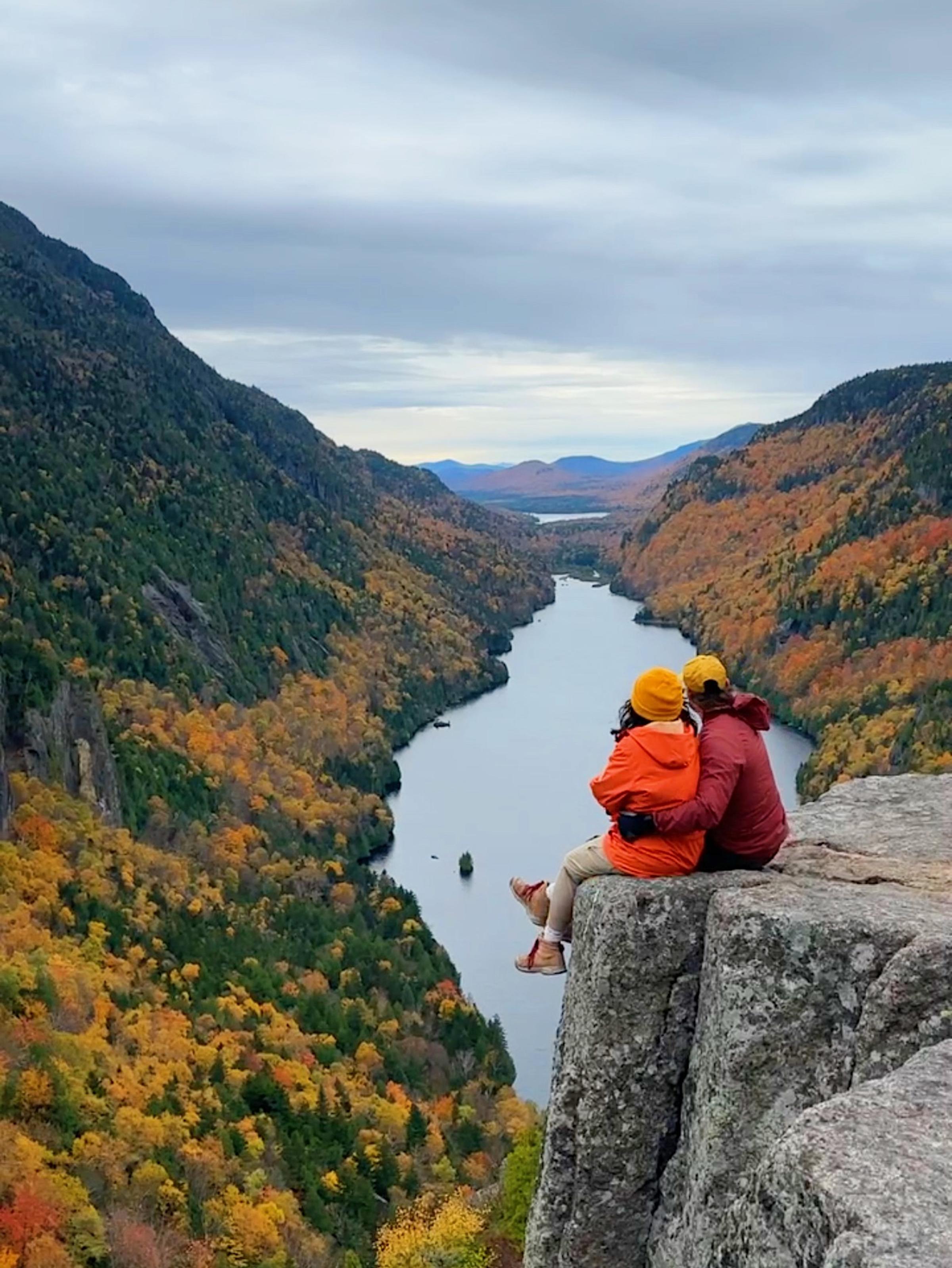 Two hikers in orange and red jackets sit together on a rocky cliff edge, embracing as they overlook a stunning river valley flanked by vibrant autumn foliage in orange, yellow, and green.