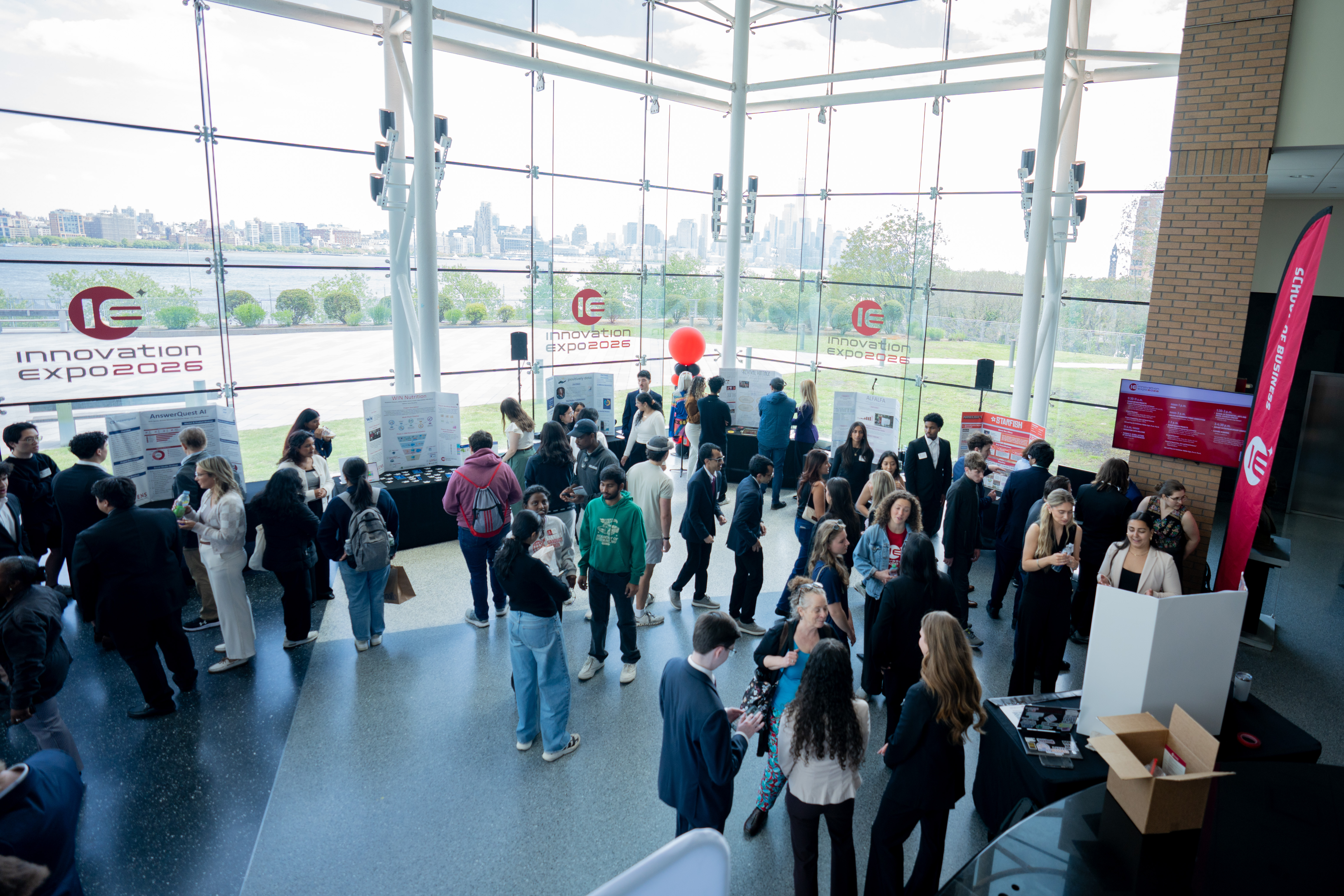 Dozens of students and attendees mingle in a bright atrium with floor-to-ceiling glass walls overlooking the NYC skyline. Innovation Expo 2026 banners and project displays are visible throughout the space, along with a red balloon marking one exhibit. 