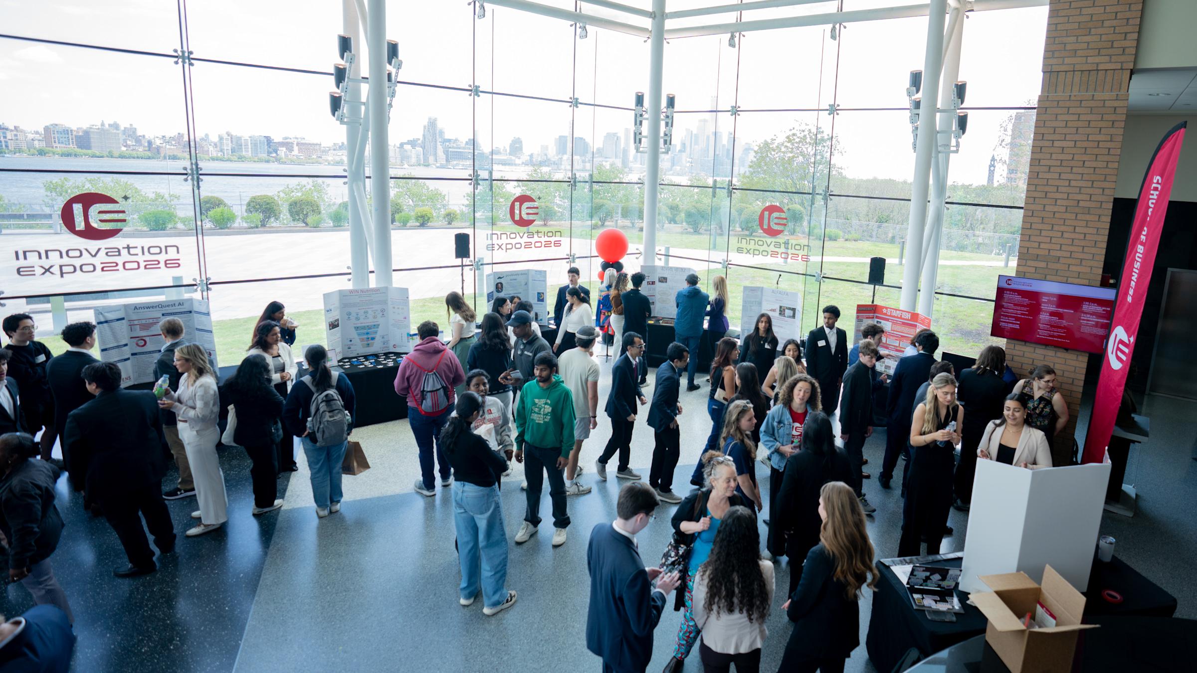 Dozens of students and attendees mingle in a bright atrium with floor-to-ceiling glass walls overlooking the NYC skyline. Innovation Expo 2026 banners and project displays are visible throughout the space, along with a red balloon marking one exhibit.