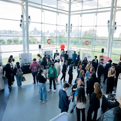 Dozens of students and attendees mingle in a bright atrium with floor-to-ceiling glass walls overlooking the NYC skyline. Innovation Expo 2026 banners and project displays are visible throughout the space, along with a red balloon marking one exhibit.