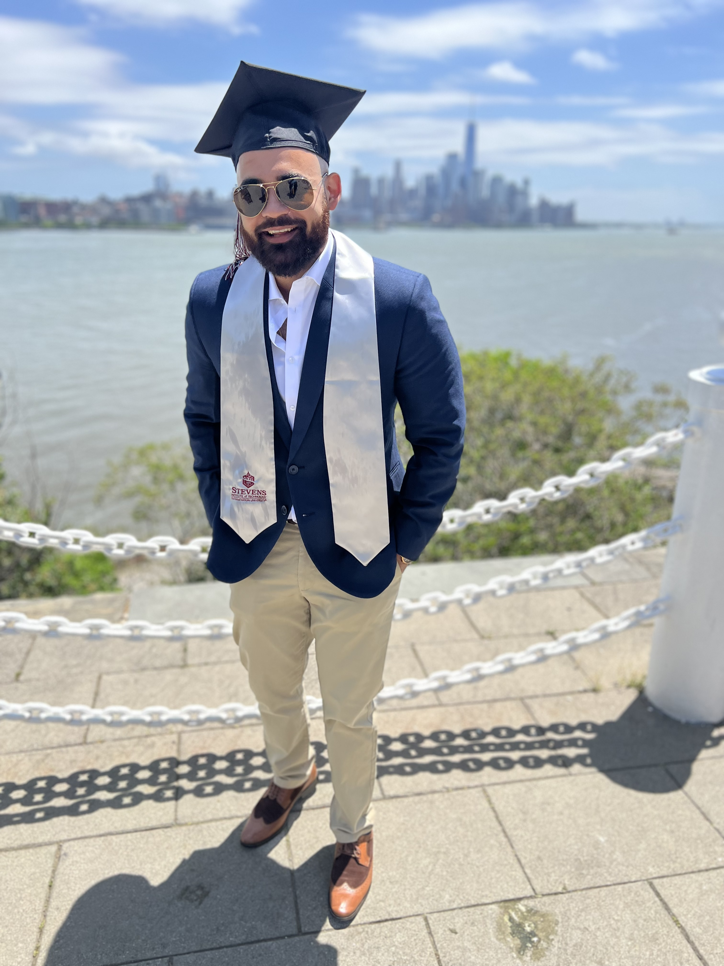 Rohan Chathrath stands with the New York City skyline in the background, wearing his graduation cap and sash.