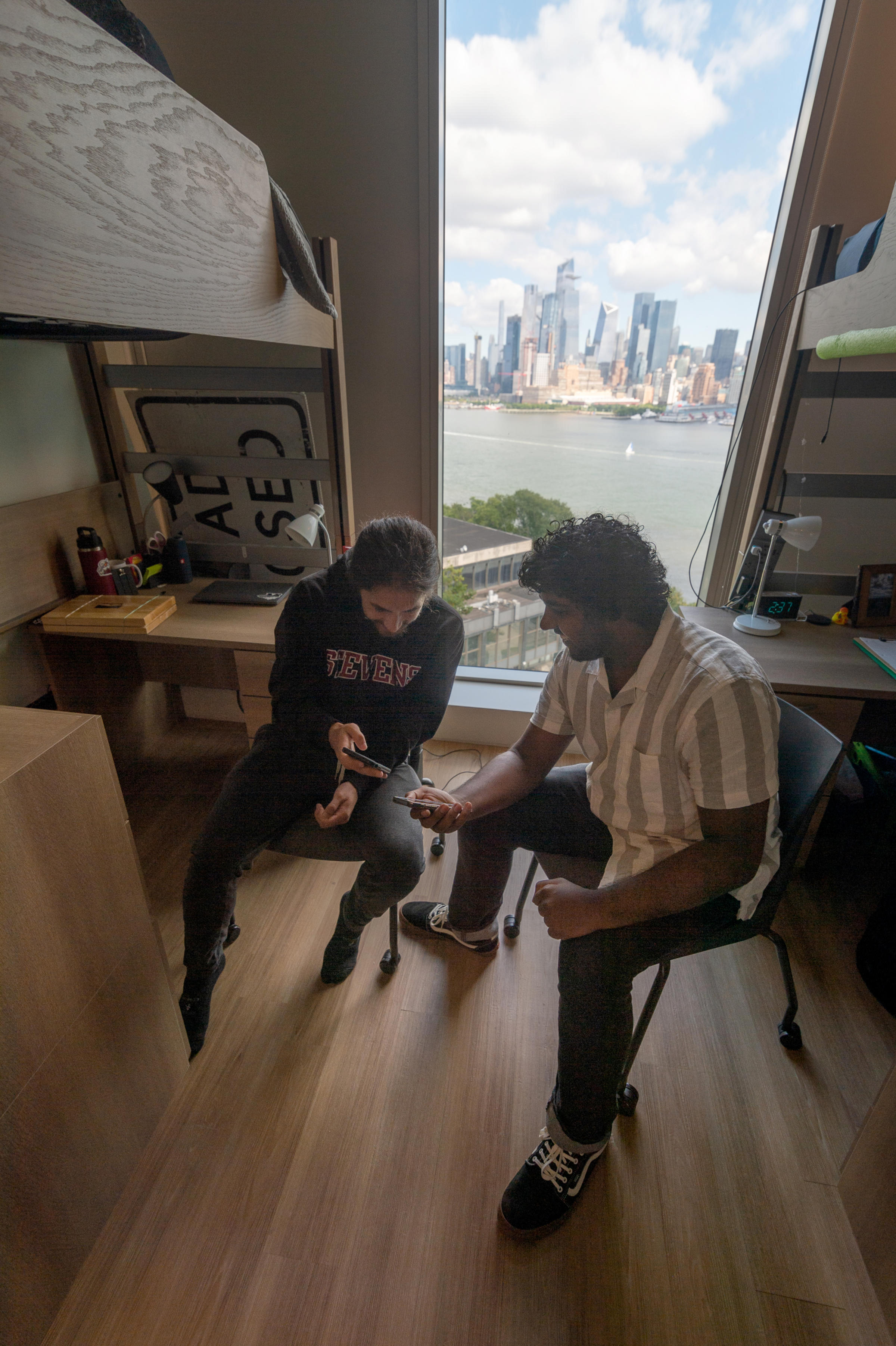 Students in dorm show each other their phones with New York city skyline through window behind them.