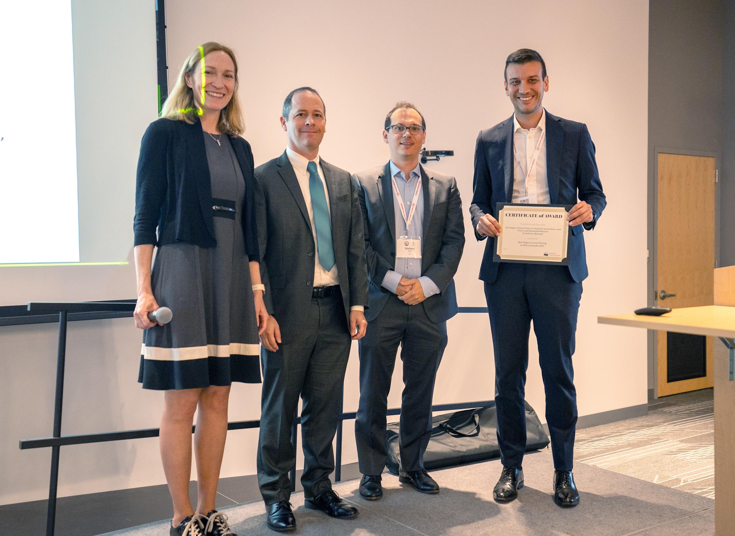 Four professionals pose for an award ceremony photo in a modern conference room. A woman in a navy dress with white stripes stands beside three men in dark business suits, with one holding a certificate, all smiling at the camera.